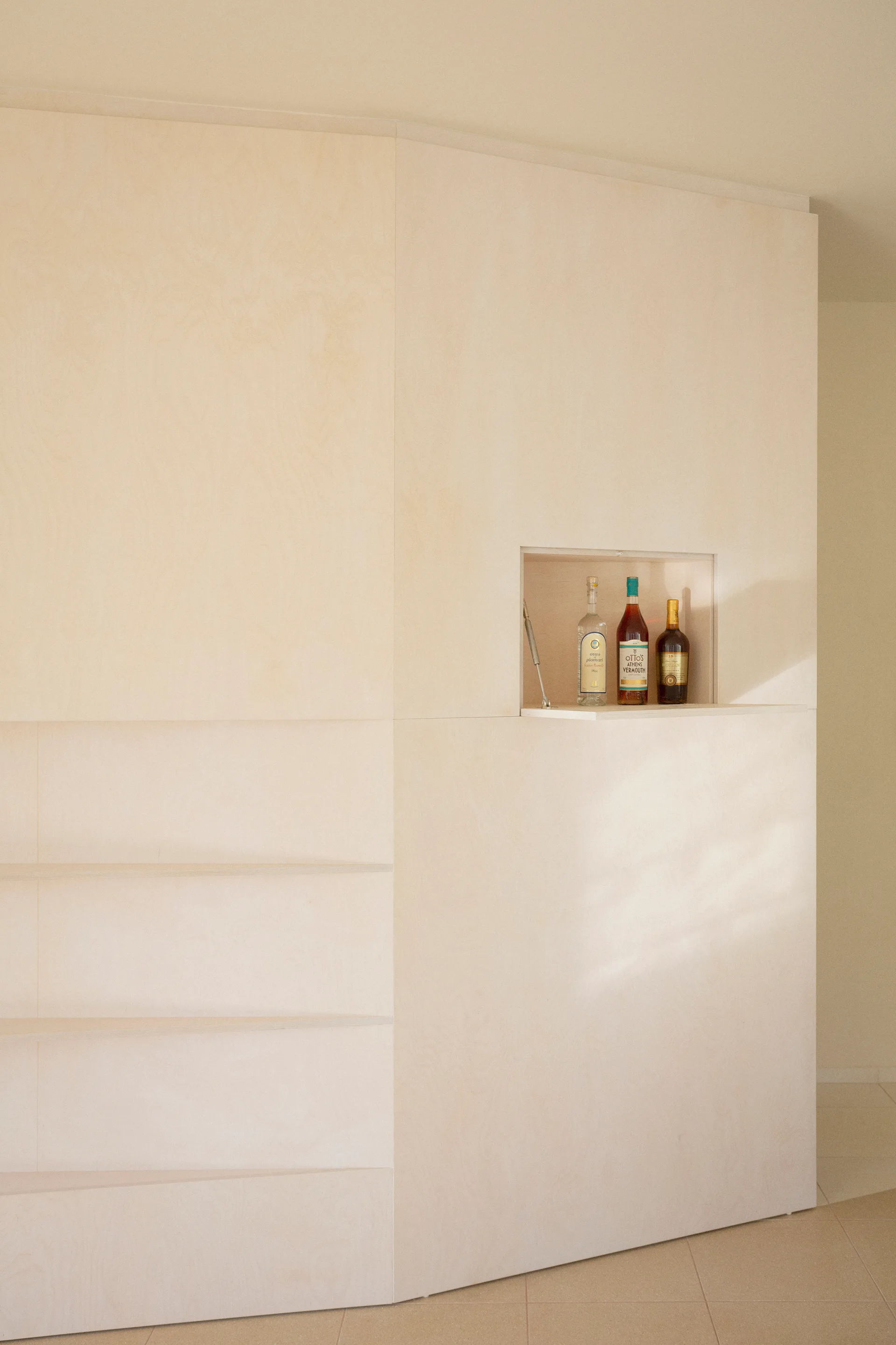 A minimalist light wood plywood cabinet wall features flat panels and a small rectangular recessed bar shelf displaying bottles.