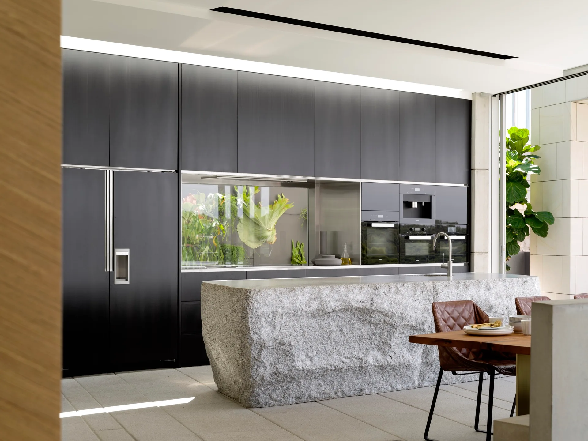 A modern kitchen featuring a textured grey stone island, flat-panel dark cabinets with integrated appliances, and a dining table with brown leather chairs.
