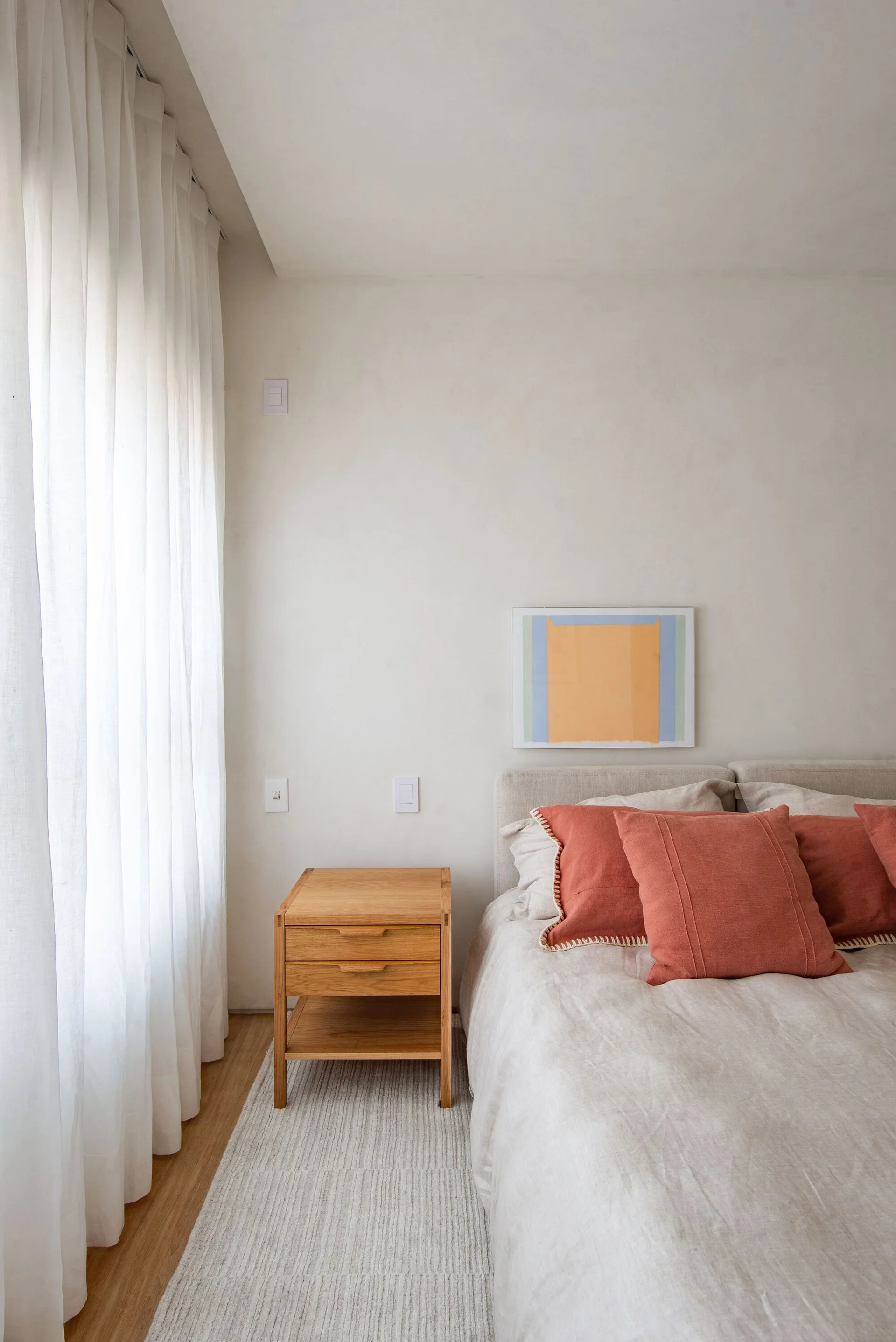 A light-filled minimalist bedroom displays a bed with terracotta linen pillows next to a light wood nightstand and sheer white curtains.