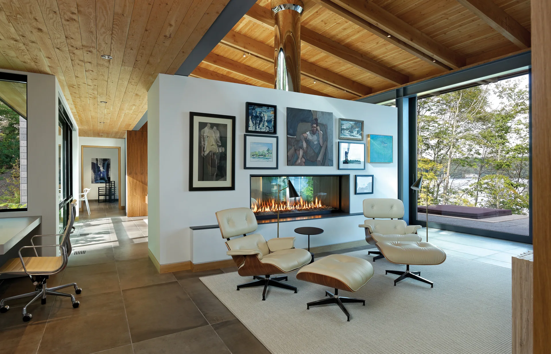 Two cream lounge chairs sit on a rug in front of a linear fireplace beneath a gallery wall in a room with a wood ceiling.