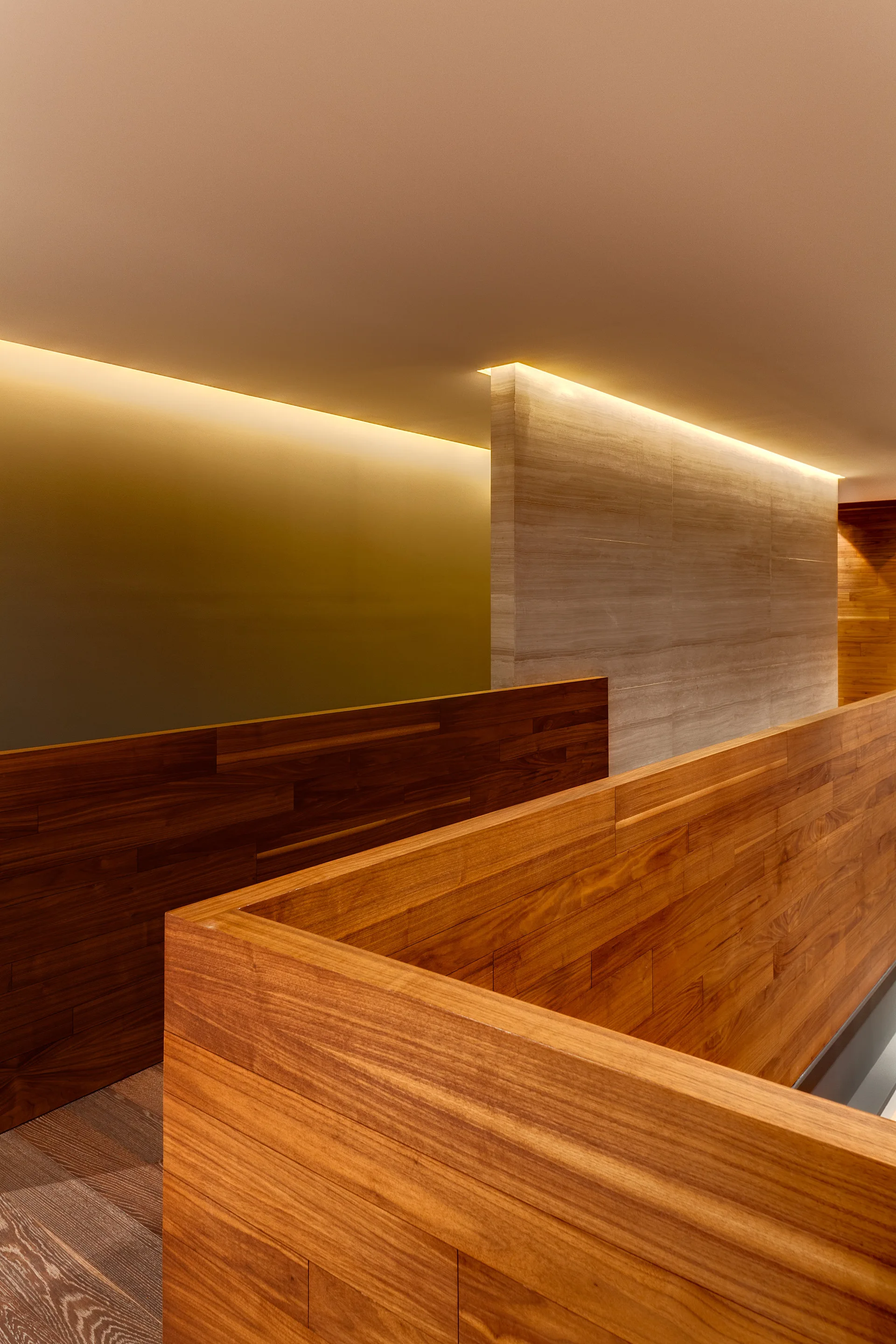 A modern corridor featuring horizontal wood paneled walls, a textured stone partition wall, and warm ambient cove lighting along the ceiling.