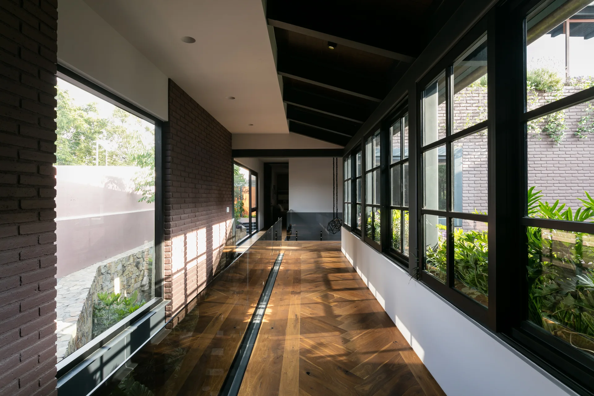 A modern hallway features herringbone patterned wood flooring, a textured brick wall, and black-framed windows overlooking outdoor greenery.
