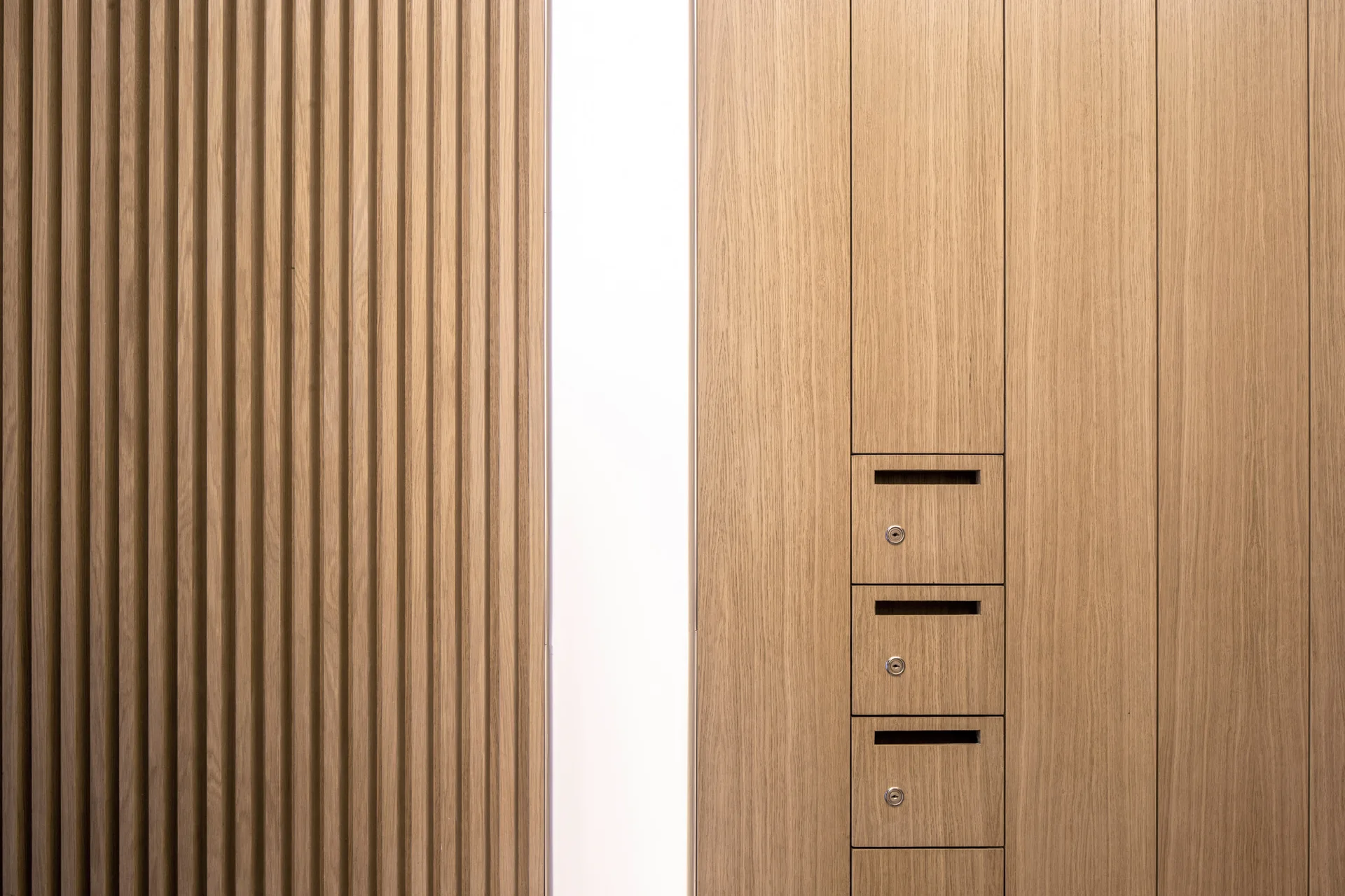 A wood paneled wall features a fluted slatted section separated by a bright vertical light strip from flat panels with integrated lockable mail slots.