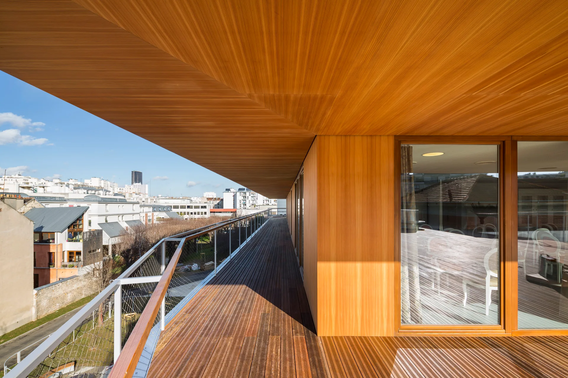 A modern outdoor terrace features warm wood cladding on the deck and ceiling, glass windows, and a metal safety railing overlooking a city.