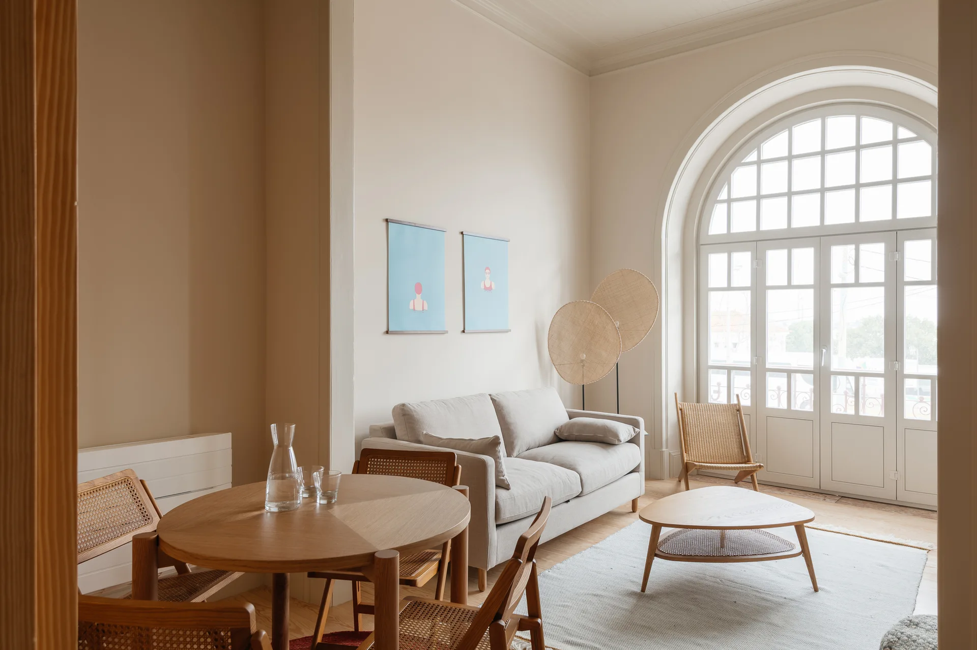 A bright living and dining room with a beige sofa, a round wooden dining table, cane chairs, and a large arched glass window.