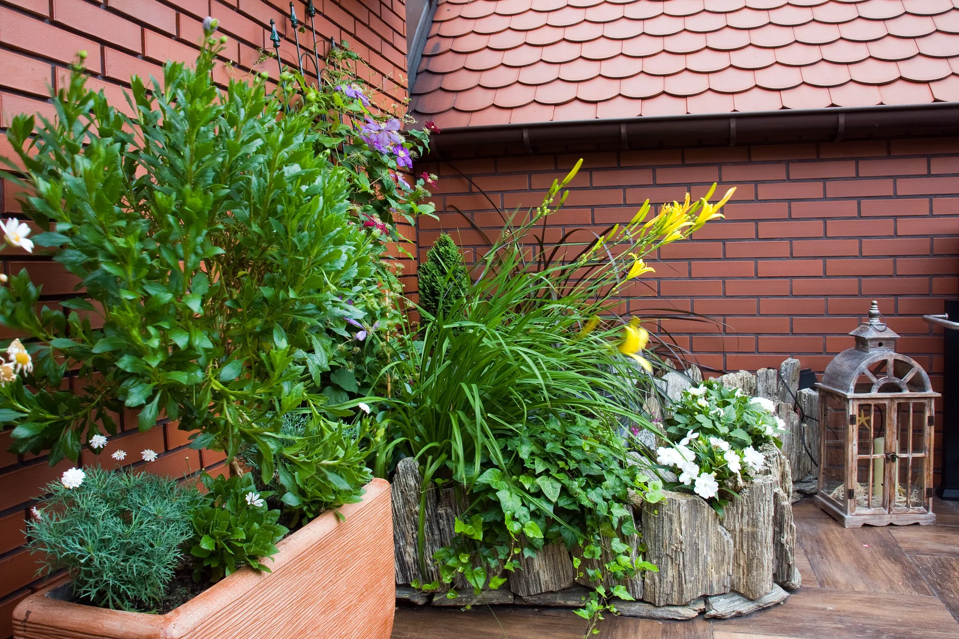 A rooftop terrace features lush green plants and yellow flowers in rustic wood planters next to a weathered lantern against a red brick wall.