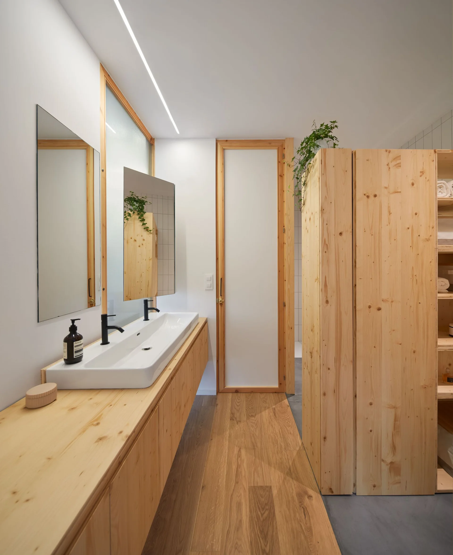 A minimalist bathroom with a light wood floating vanity, double white sinks, matte black faucets, and tall pine storage cabinets.