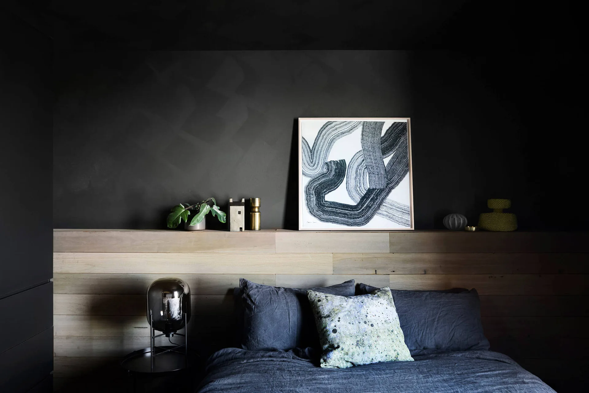 A modern bedroom features a dark gray wall, horizontal wood paneling used as a headboard, a display ledge with abstract art, and dark linen bedding.