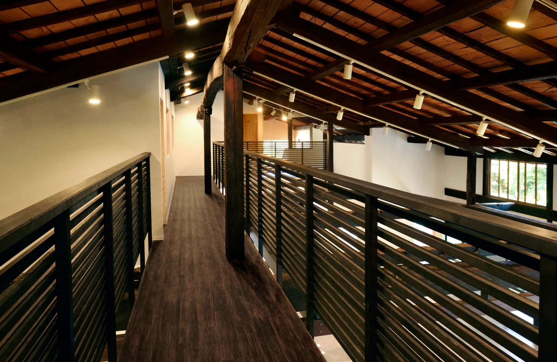 An elevated dark wood mezzanine walkway features horizontal slatted railings beneath a pitched roof with exposed timber beams and track lighting.