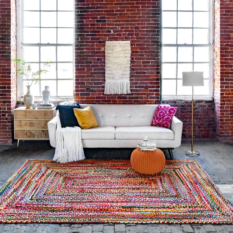 A light grey sofa and a multicolored braided rug sit against an exposed red brick wall with large industrial windows.
