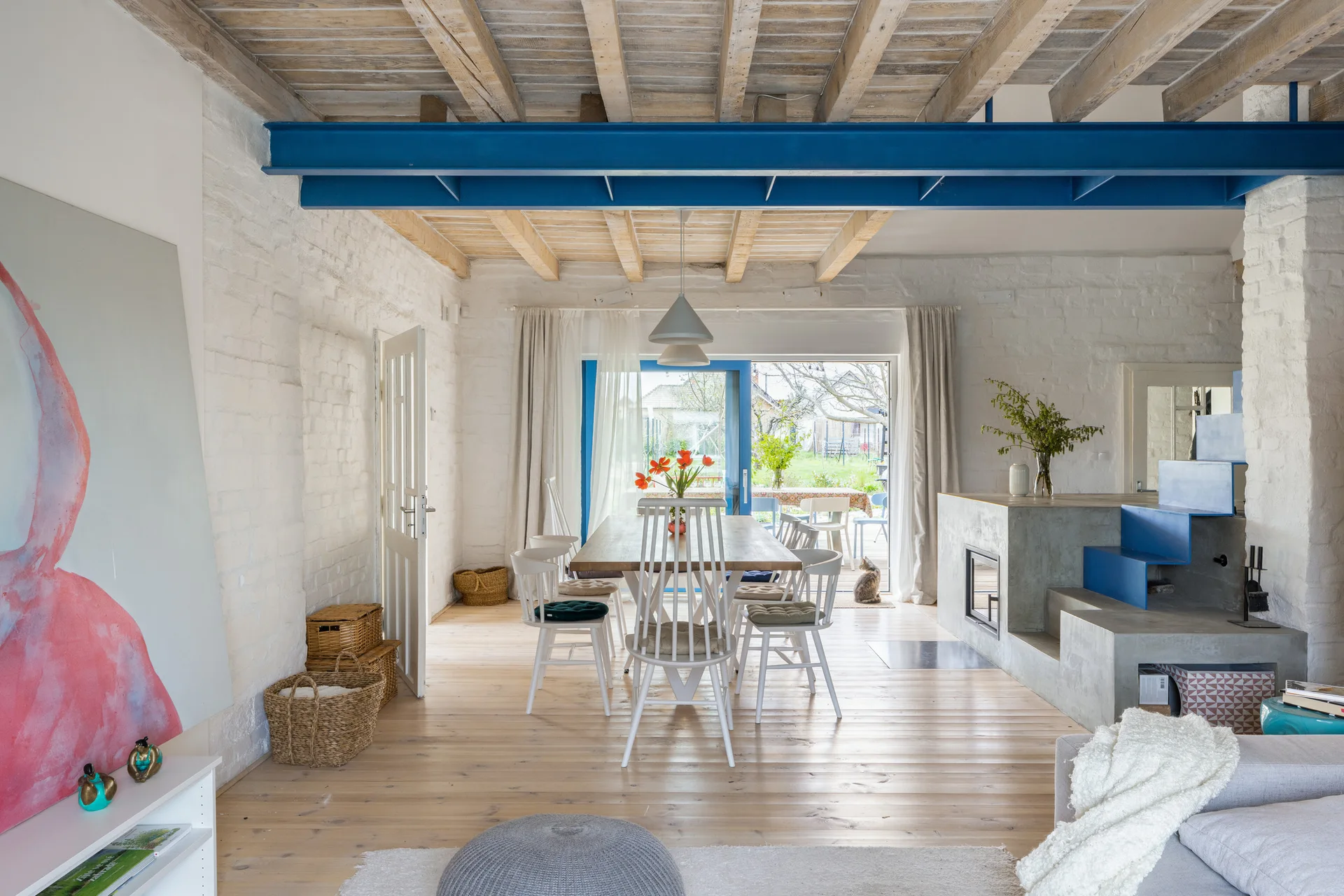An open dining room features a rustic wood table and white spindle chairs underneath a raw wood ceiling accented by a painted blue steel beam.