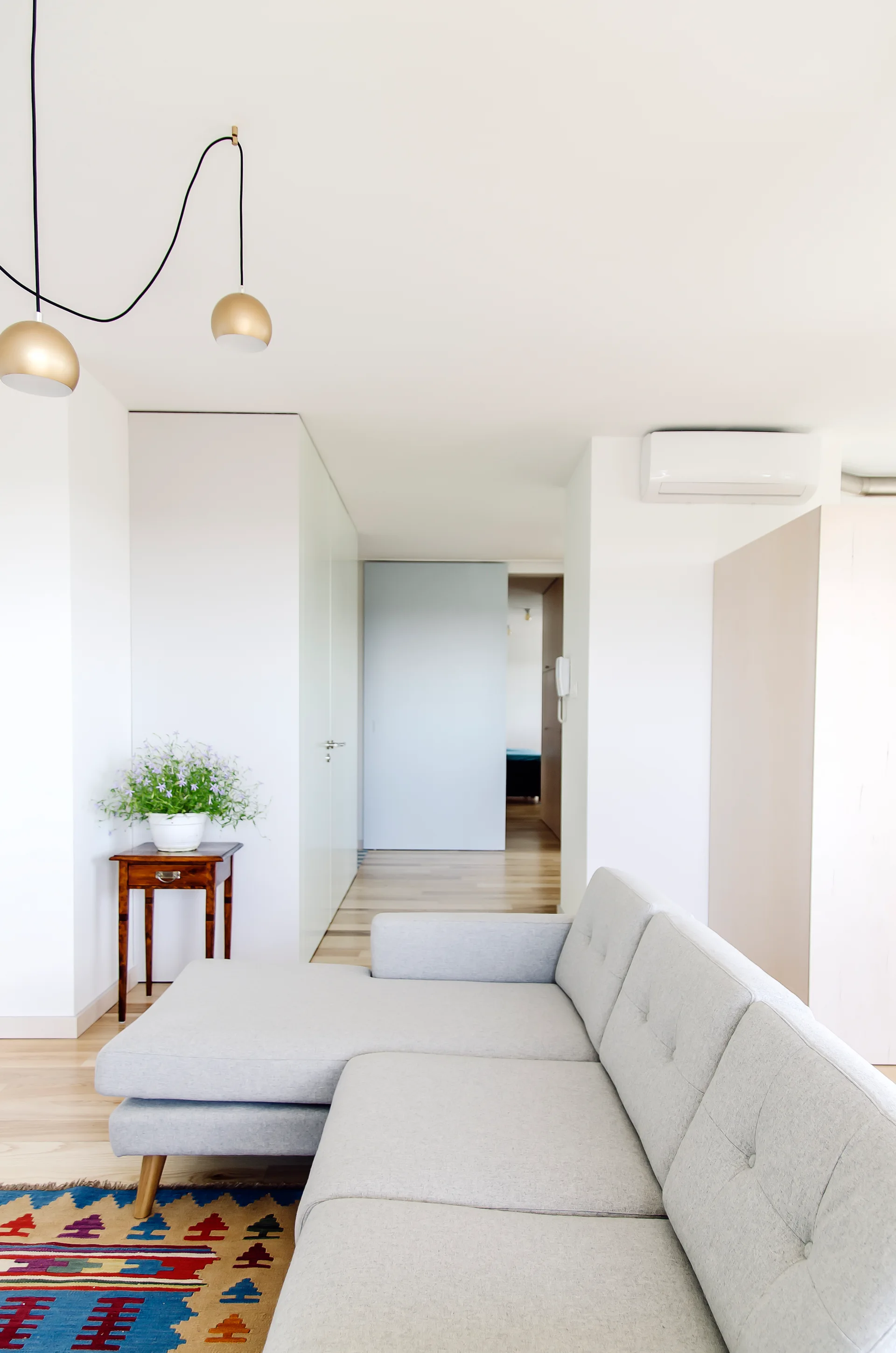 A light grey sectional sofa sits on a colorful geometric patterned rug in a minimalist white living room with light wood floors and a brass pendant light.