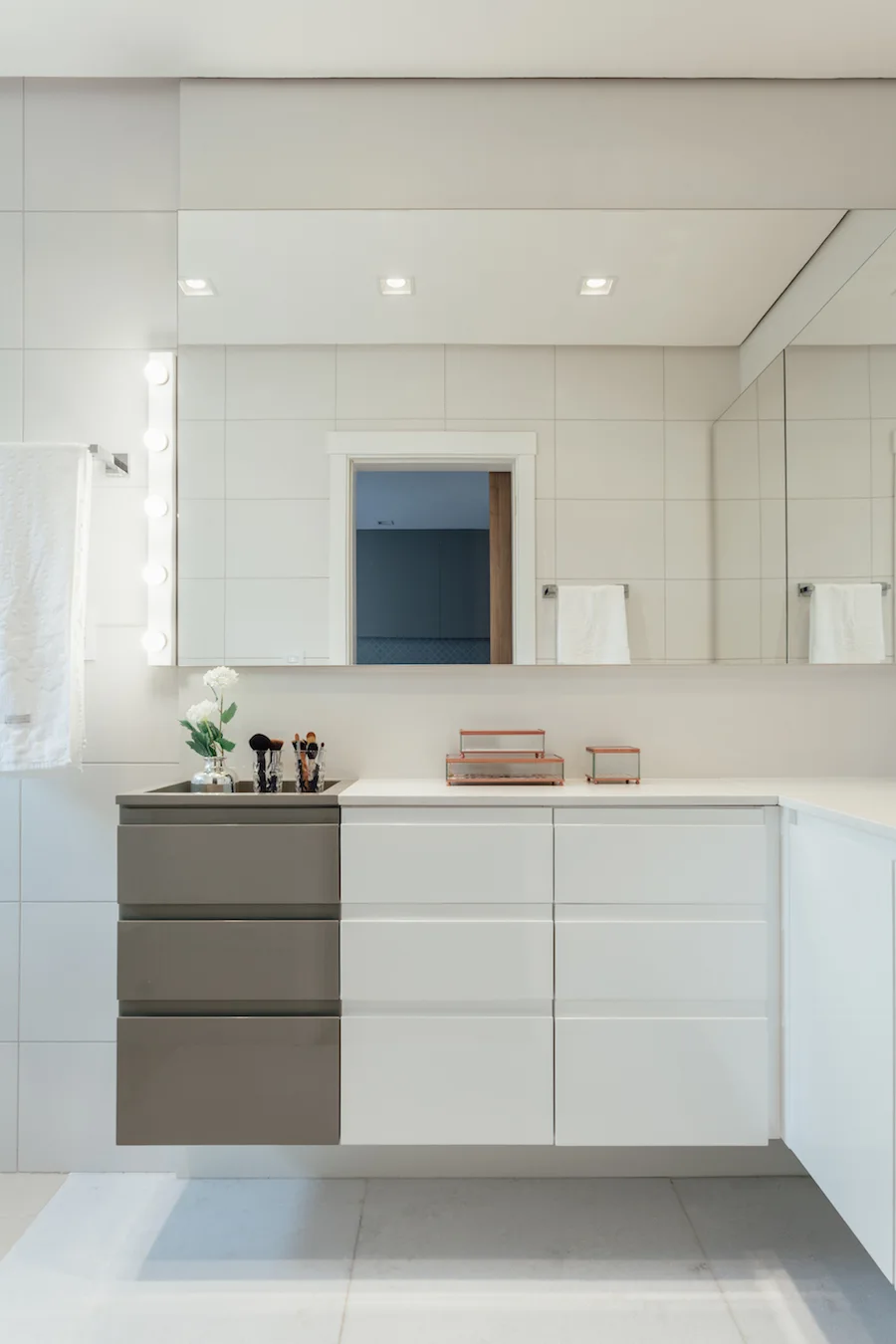 A modern bathroom vanity with white and grey glossy drawers is set against a wall of large white tiles next to a light strip.
