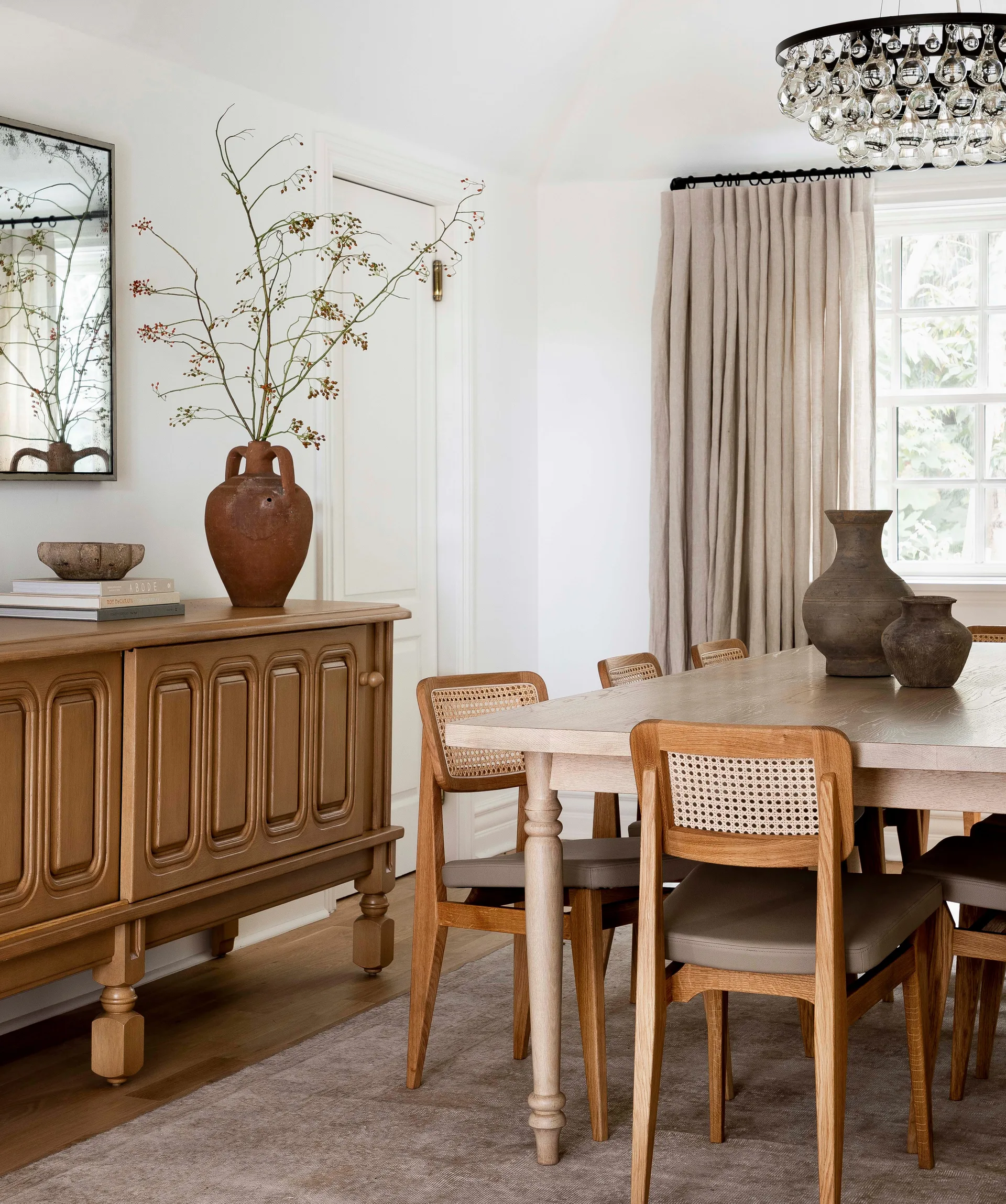 A bright dining room features a wooden table with cane-back chairs, a detailed wood sideboard, and a neutral drapery panel.