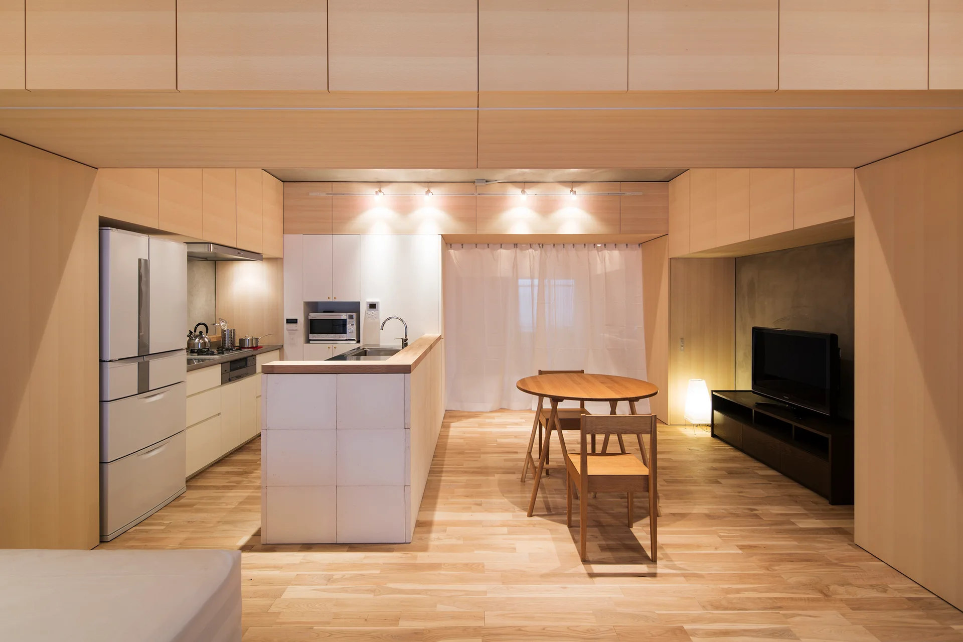 A minimalist open-concept kitchen and dining area with extensive light wood paneling, a white central island, a round dining table, and a television.