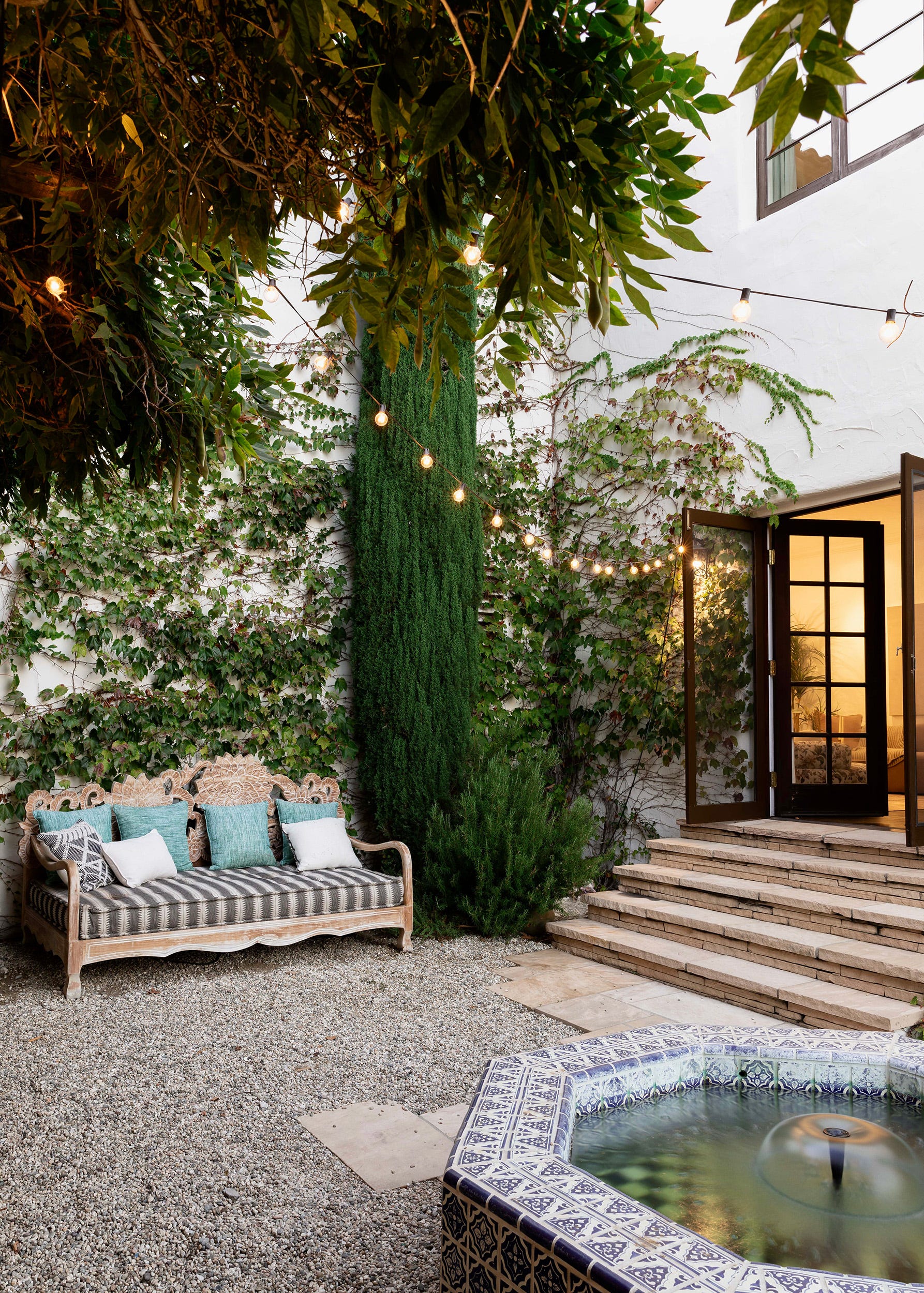 An outdoor gravel courtyard features a carved wood bench with teal pillows, an ivy-covered wall, and a blue and white tiled fountain.