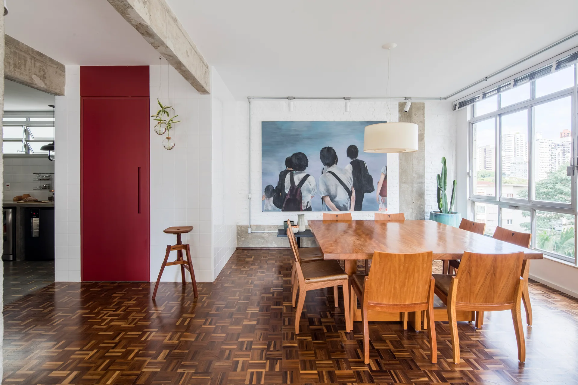 A bright dining room featuring a large wooden table and chairs, parquet wood flooring, and a bold red architectural panel wall.