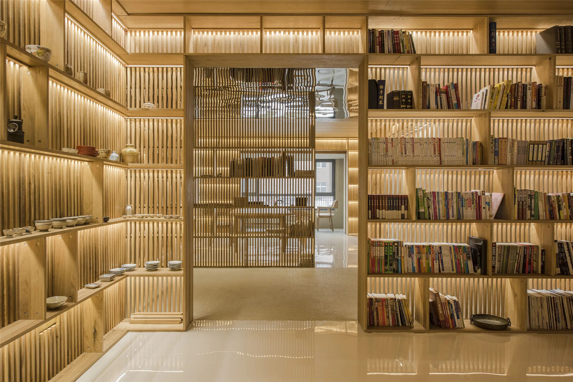 Floor-to-ceiling wooden bookshelves with integrated warm lighting surround a central doorway with a vertical wood slat screen and highly polished light floors.
