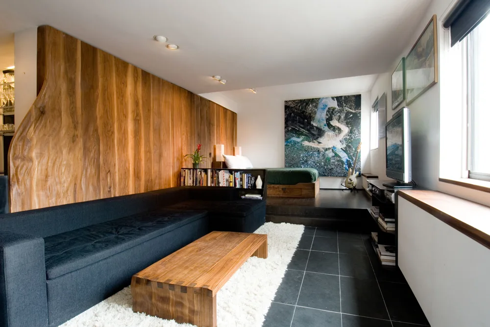 A split-level studio apartment featuring a black sectional sofa on a white shag rug, a wood-paneled accent wall, and an elevated platform bed area.