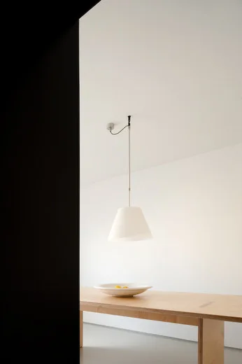 A minimalist dining room featuring a light wood table, a white cone-shaped pendant light suspended from the ceiling, and a white decorative bowl.