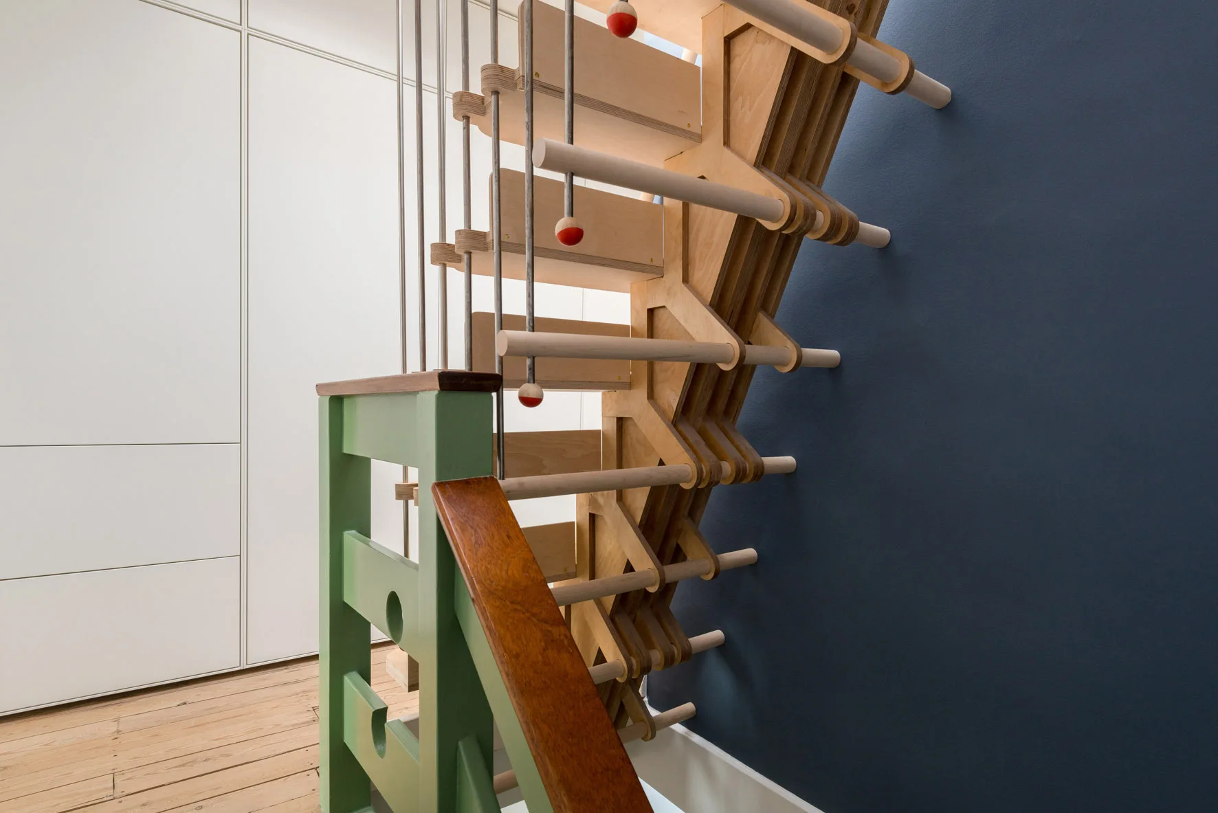 A custom light wood staircase featuring unique geometric joinery stands between a white cabinet wall and a dark blue painted wall.