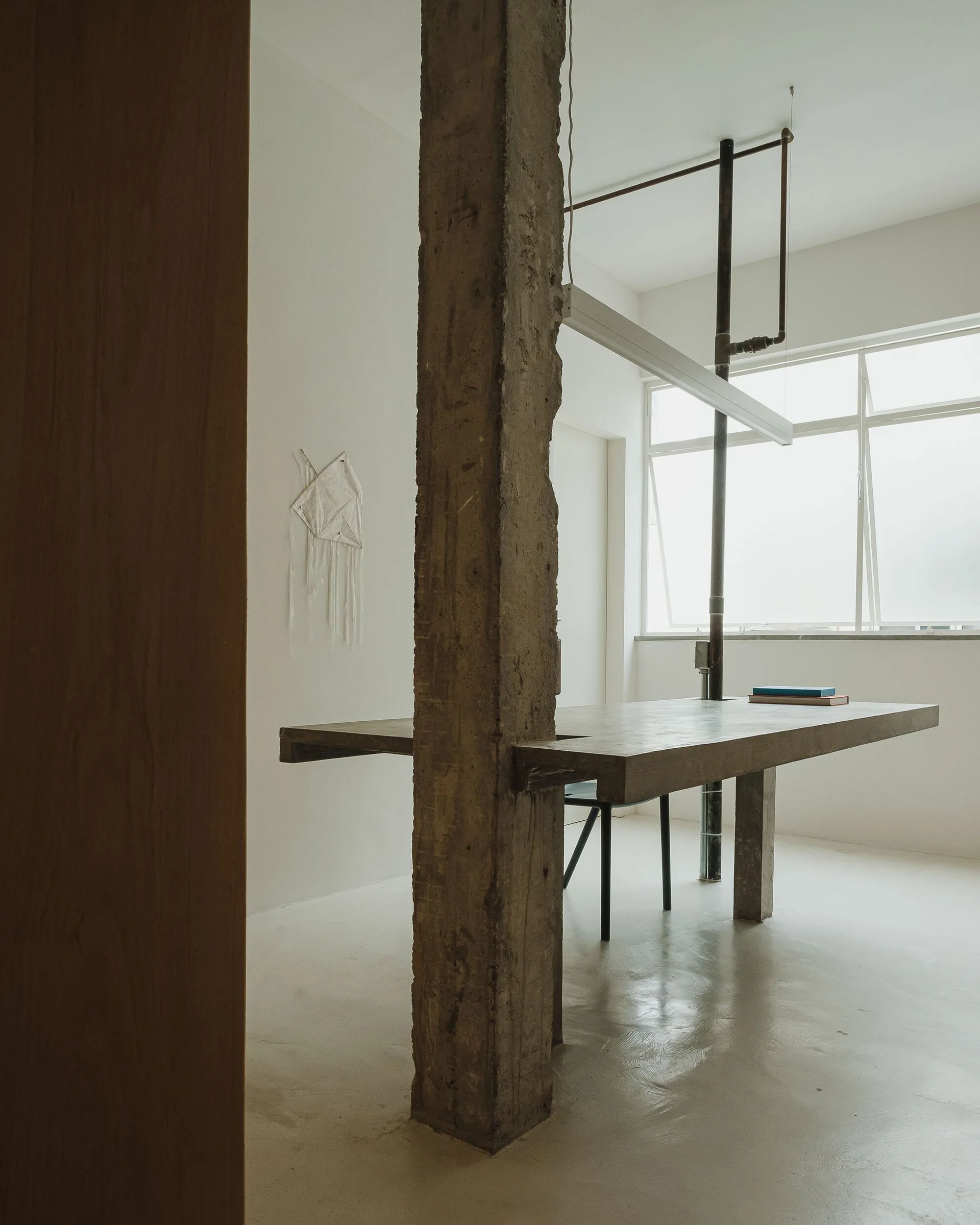 A long grey concrete table is bisected by a rough structural column in a bright minimalist room with a large window and seamless floors.