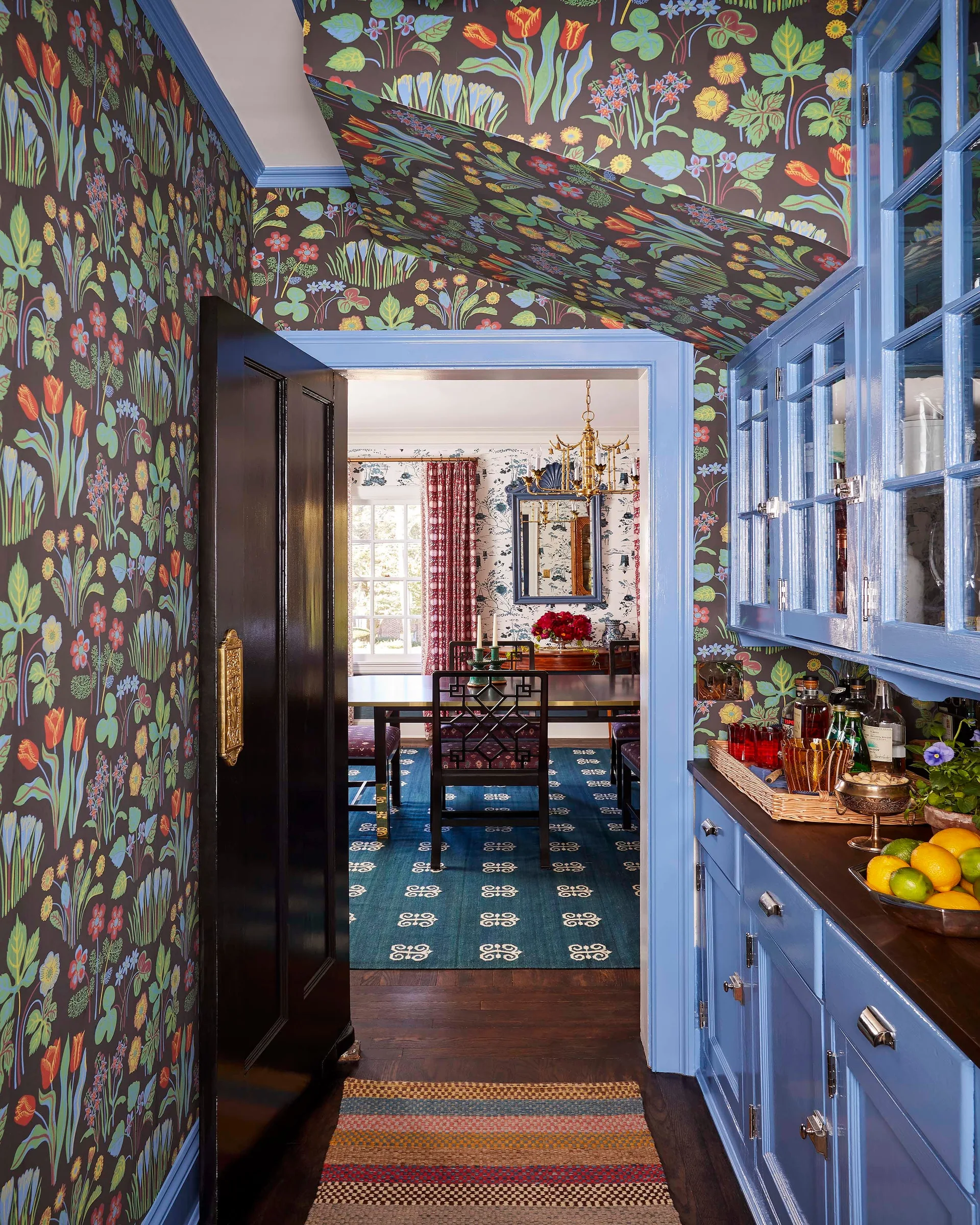 A butler's pantry features blue cabinets and dark floral wallpaper that extends to the ceiling, looking into a patterned dining room.