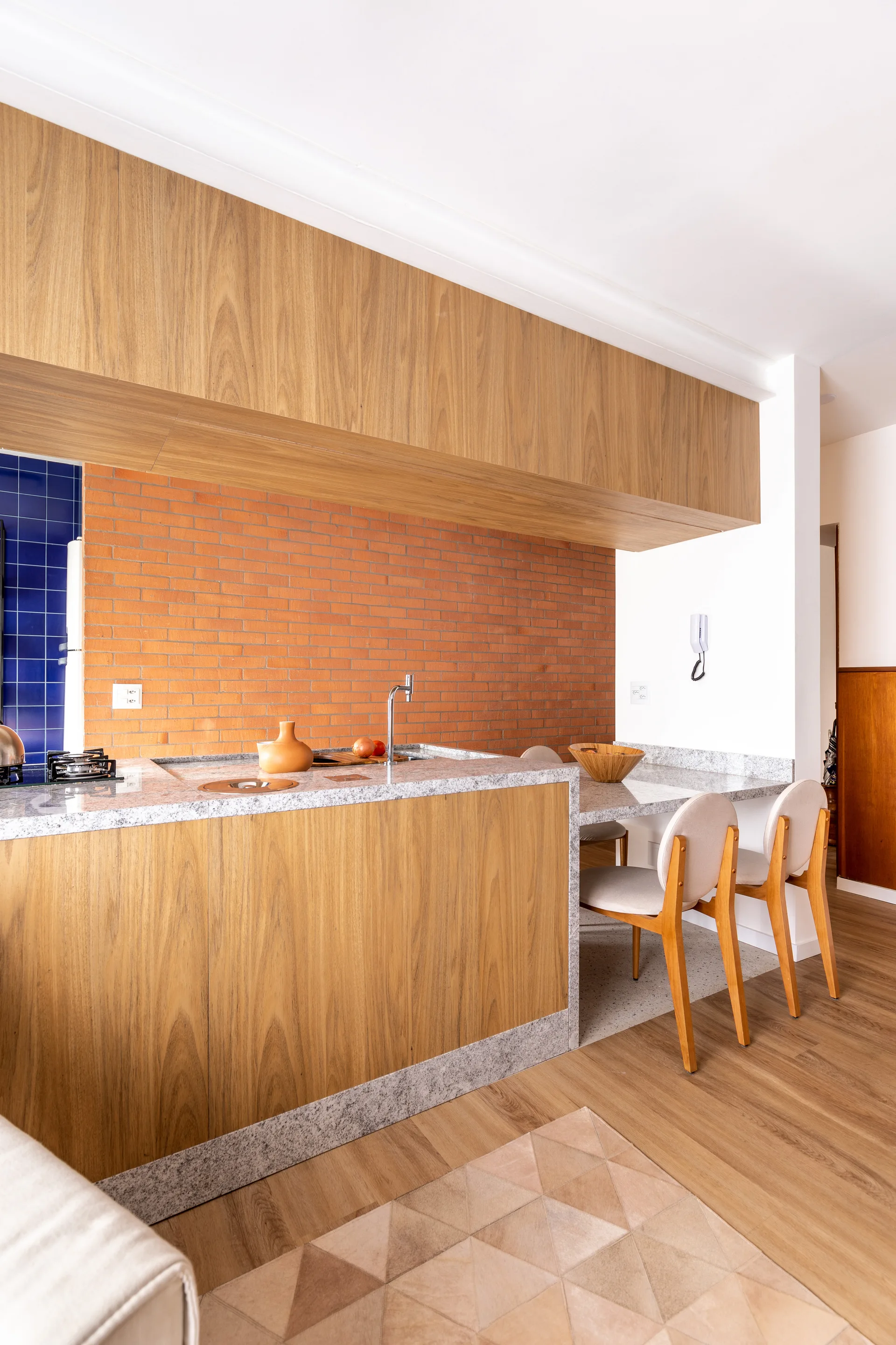 A modern kitchen featuring a wood-paneled island with grey stone countertops, a breakfast bar with two chairs, and an exposed red brick backsplash.