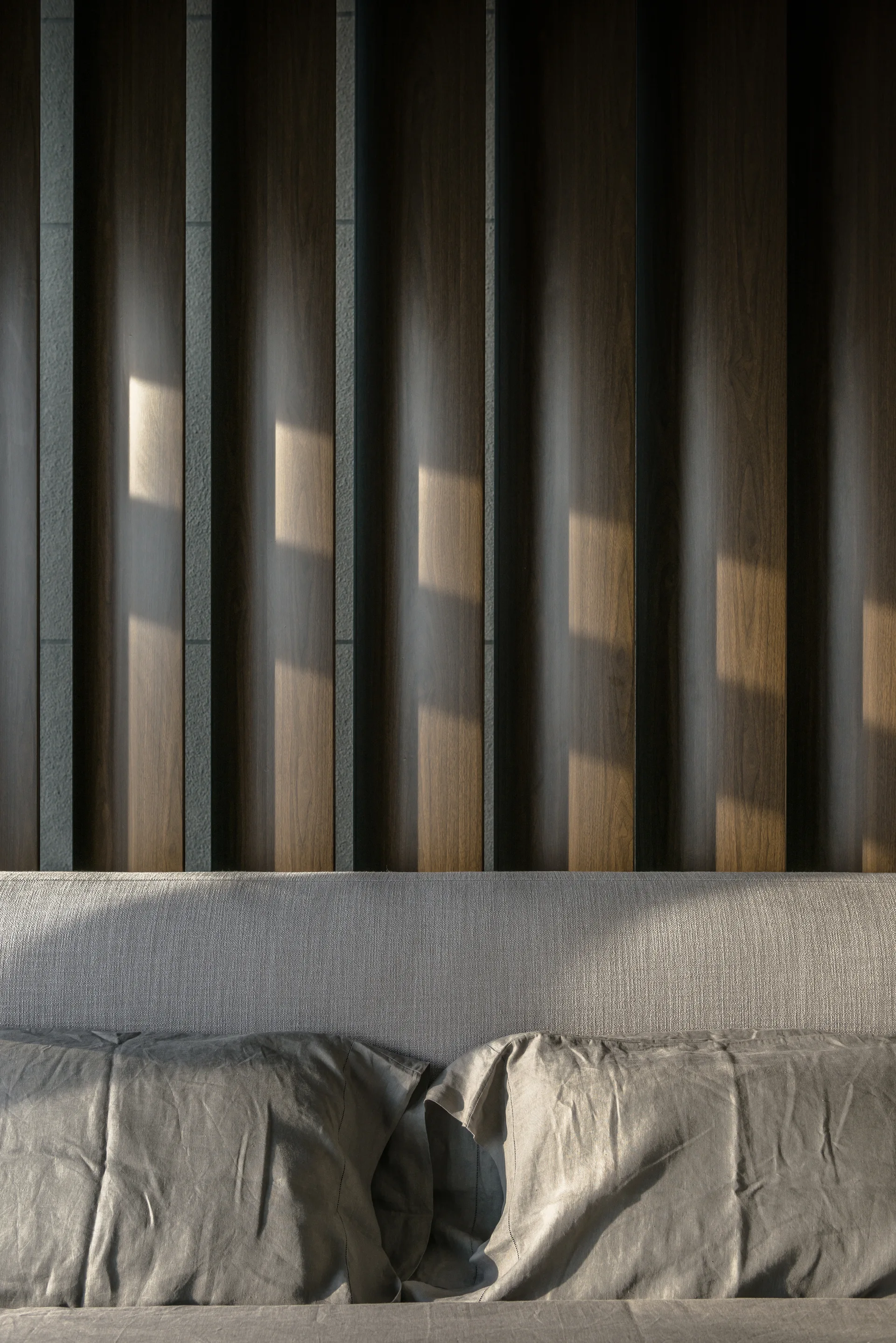 A close-up view of a grey upholstered headboard and matching linen pillows set against a textured wall of vertical dark wood panels.