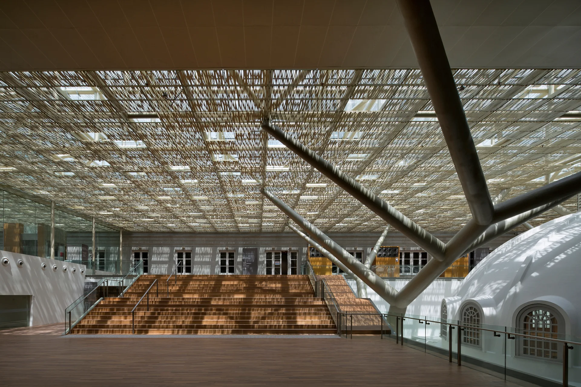 A spacious indoor atrium features a wide wooden staircase under a large metal branched pillar and an intricate overhead wood latticework ceiling.
