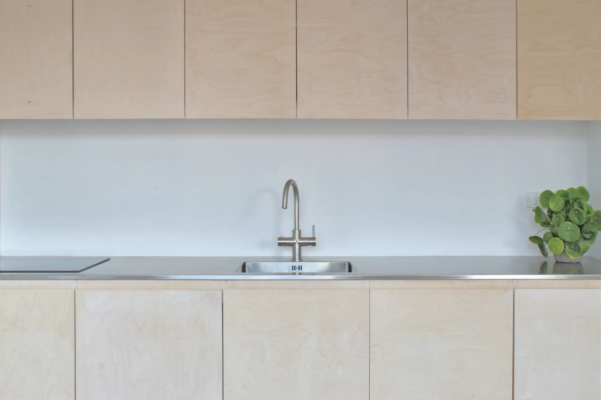 Modern kitchen featuring light-colored plywood cabinets, a stainless steel countertop with an integrated sink, and a white backsplash.