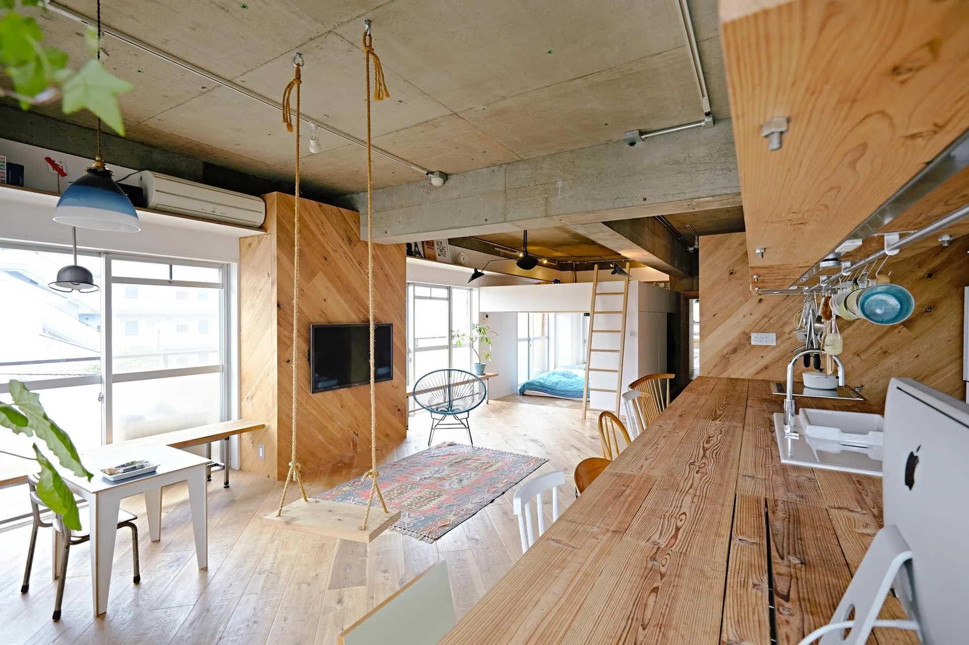 An open-plan loft apartment featuring an exposed concrete ceiling, diagonal wood paneling on the columns, an indoor rope swing, and a long wooden counter.