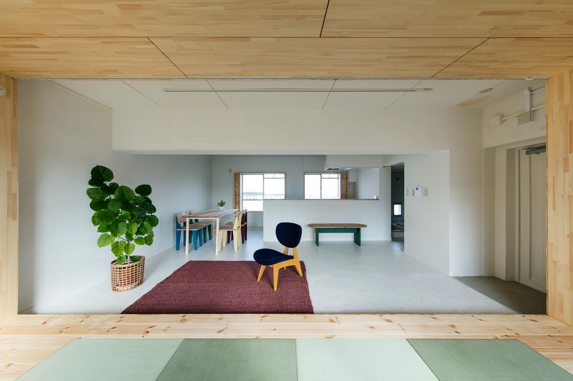 An open-plan living and dining area viewed from a tatami mat room, featuring a wood paneled ceiling, a burgundy rug, and minimalist furniture.