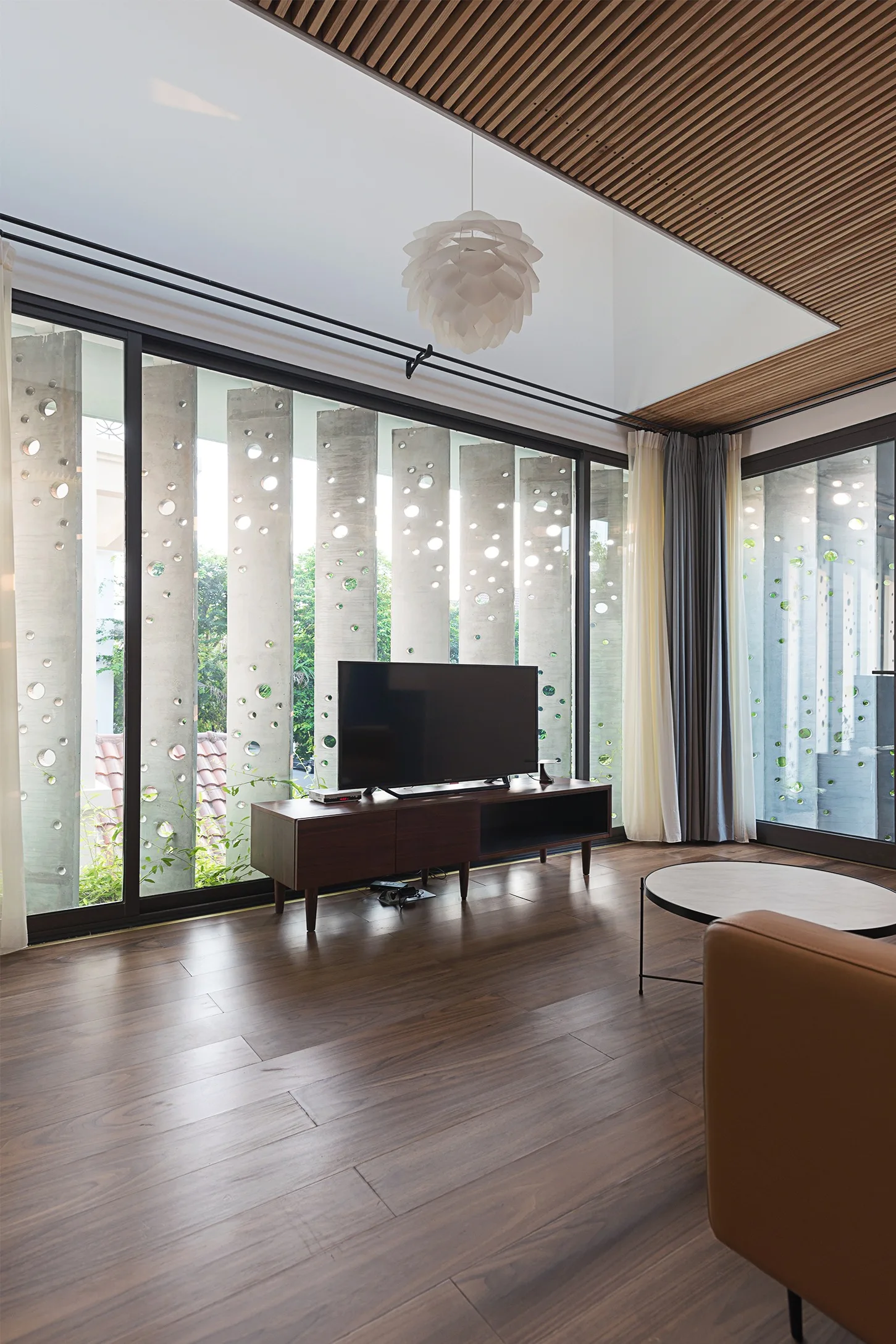 A modern living room with wood flooring, a slatted ceiling, a TV console, and floor-to-ceiling windows showing perforated concrete screens outside.