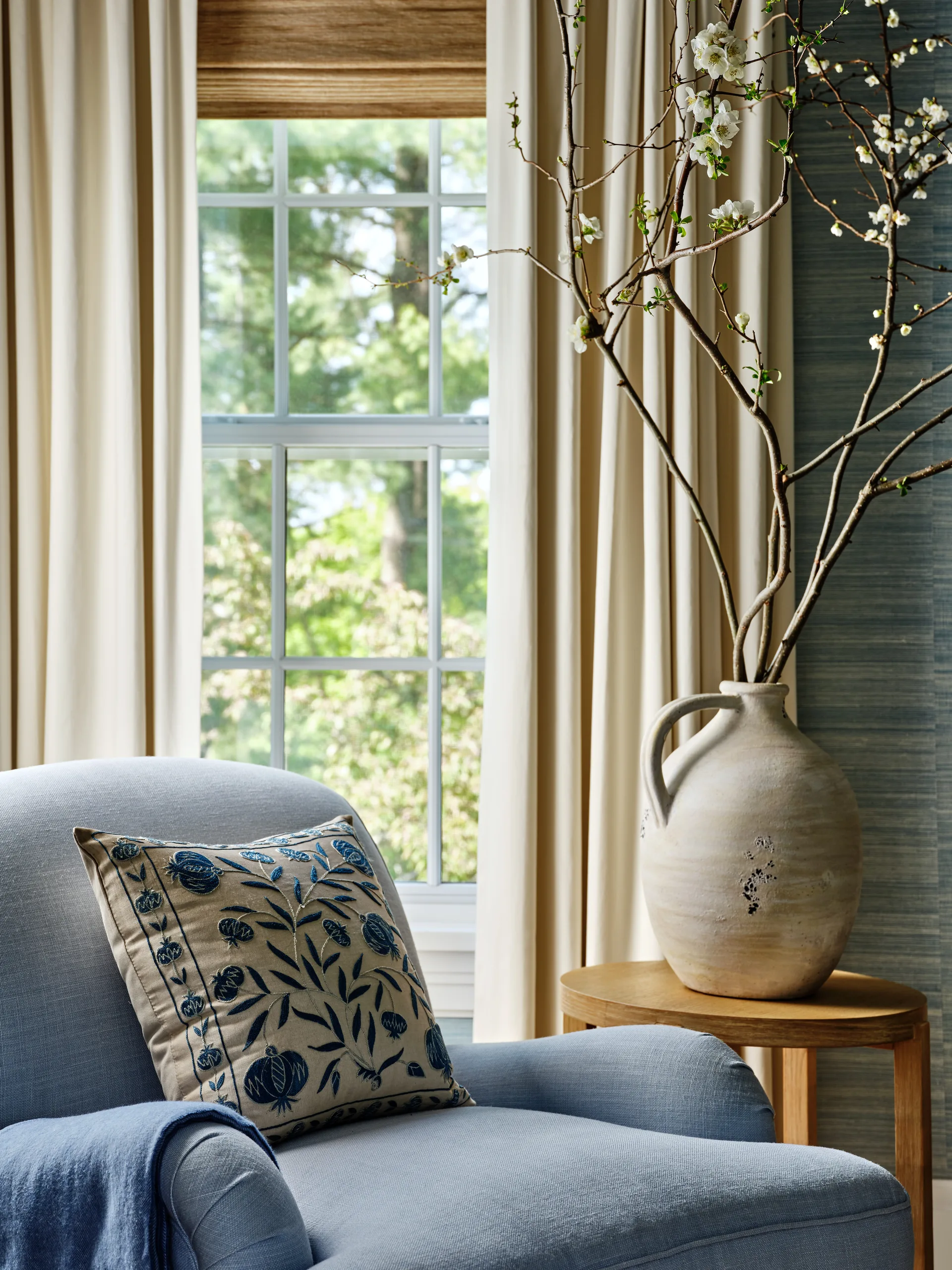 A light blue upholstered armchair with an embroidered floral pillow sits next to a wooden side table holding a ceramic jug filled with flowering branches.