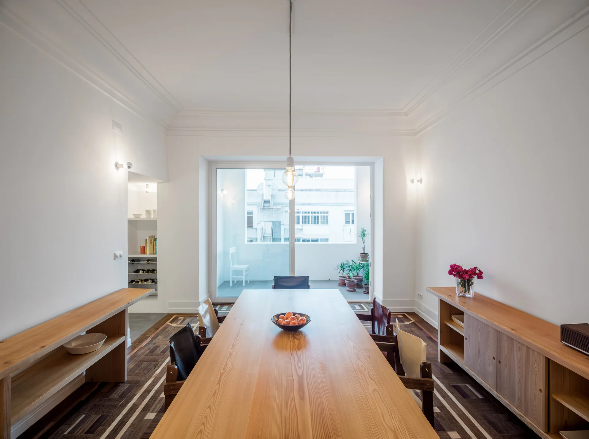 A long light wood dining table with chairs sits on an intricate parquet floor in a bright white room with sideboards and a sliding balcony door.