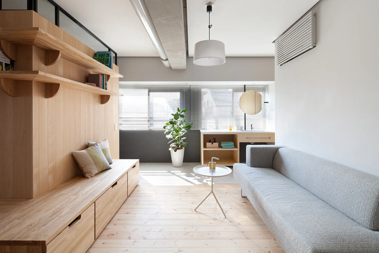 A bright living room features a custom light wood wall unit with integrated bench seating opposite a modern grey fabric sofa on wood plank flooring.