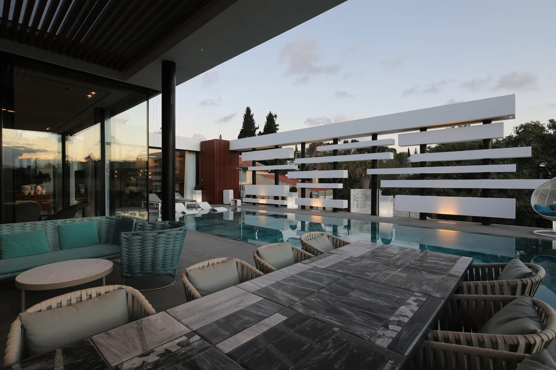 A modern outdoor terrace features a stone-topped dining table, woven chairs, a swimming pool, and a white architectural screen.
