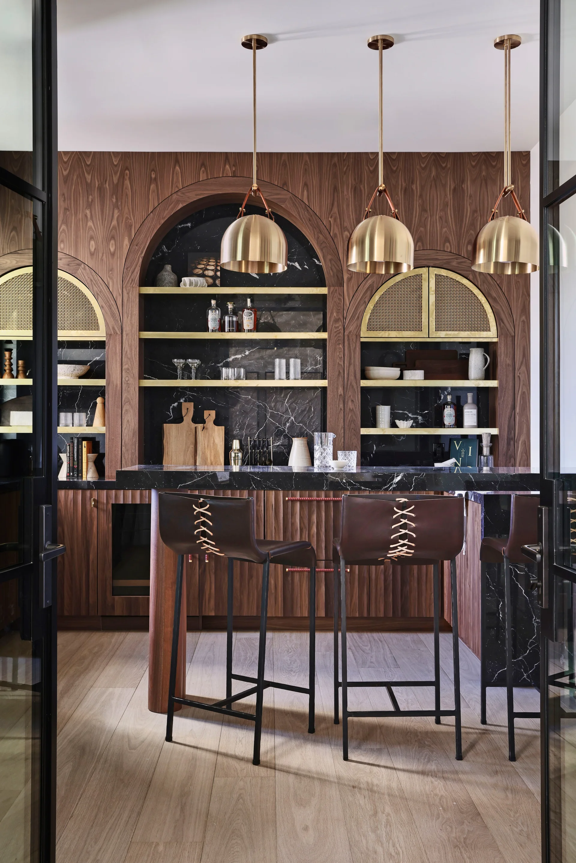 A home bar featuring arched walnut cabinetry, black marble countertops, brass dome pendant lights, and leather-backed stools with lace-up details.