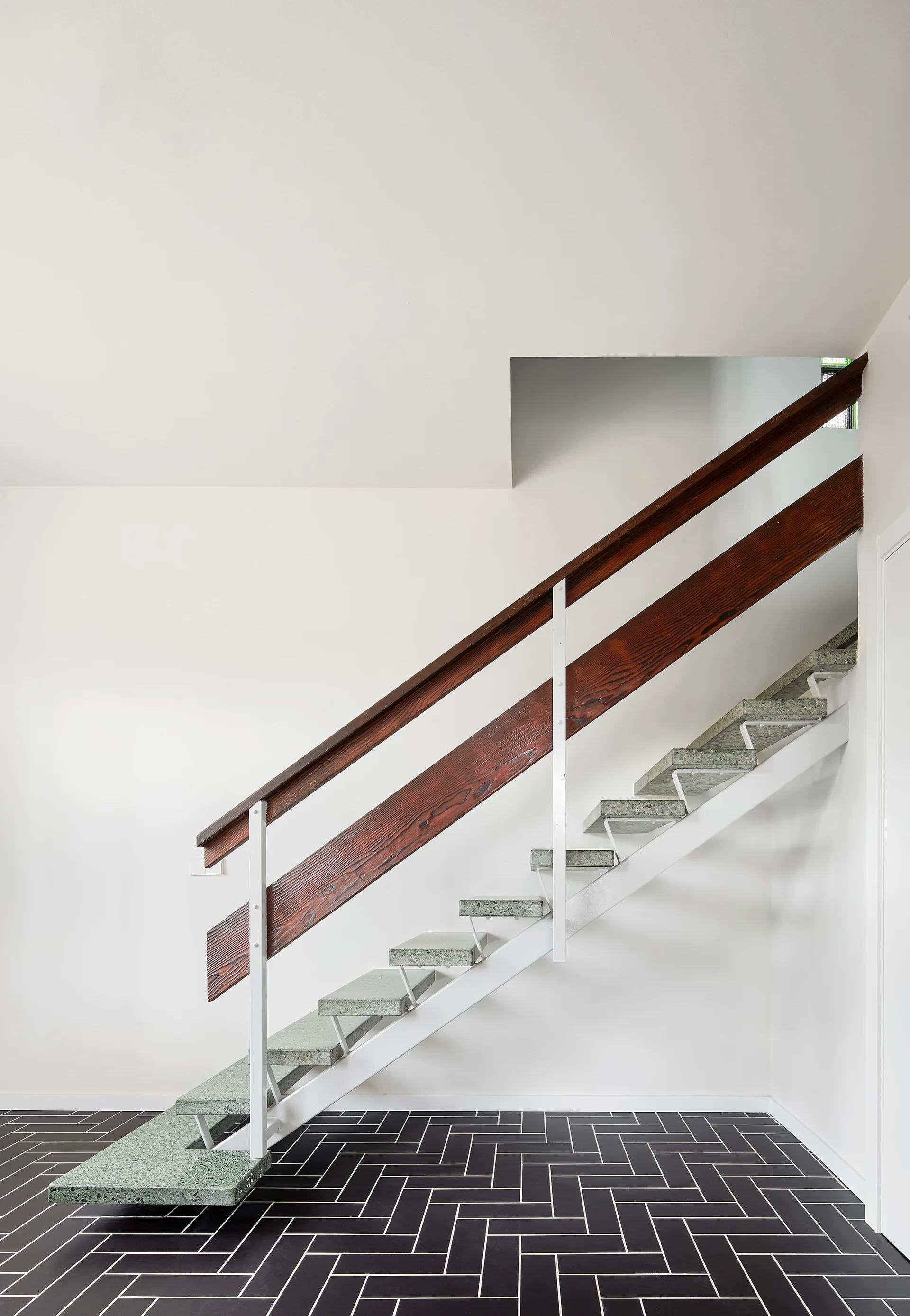A modern staircase with light green stone treads and a dark wood handrail rises above a black tiled floor laid in a herringbone pattern.