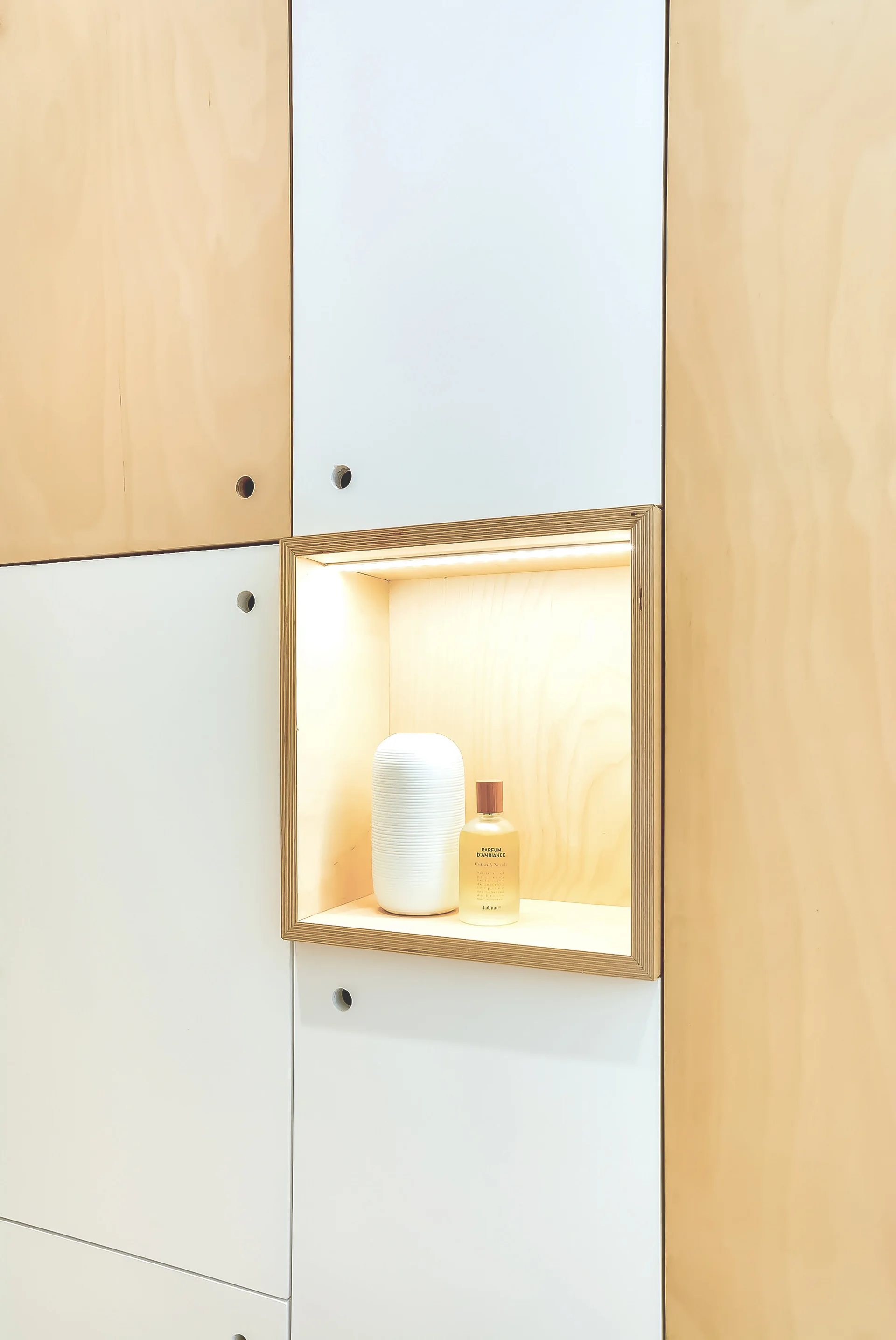 A close-up of custom wood and white cabinet panels featuring an illuminated square niche displaying a small white vase and a glass bottle.