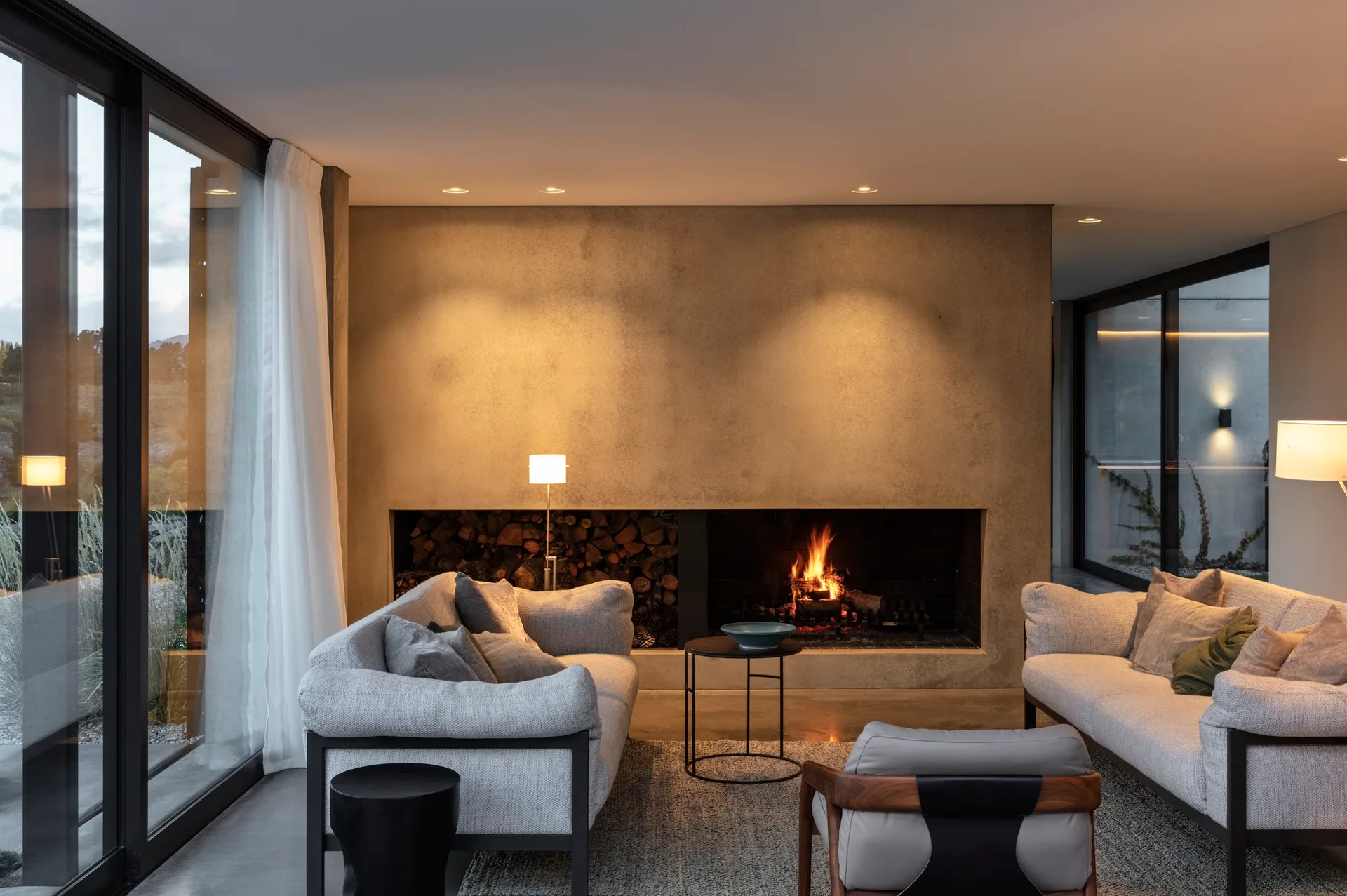 A modern living room features two light grey sofas flanking a central fireplace with an integrated wood storage niche beneath a textured plaster wall.