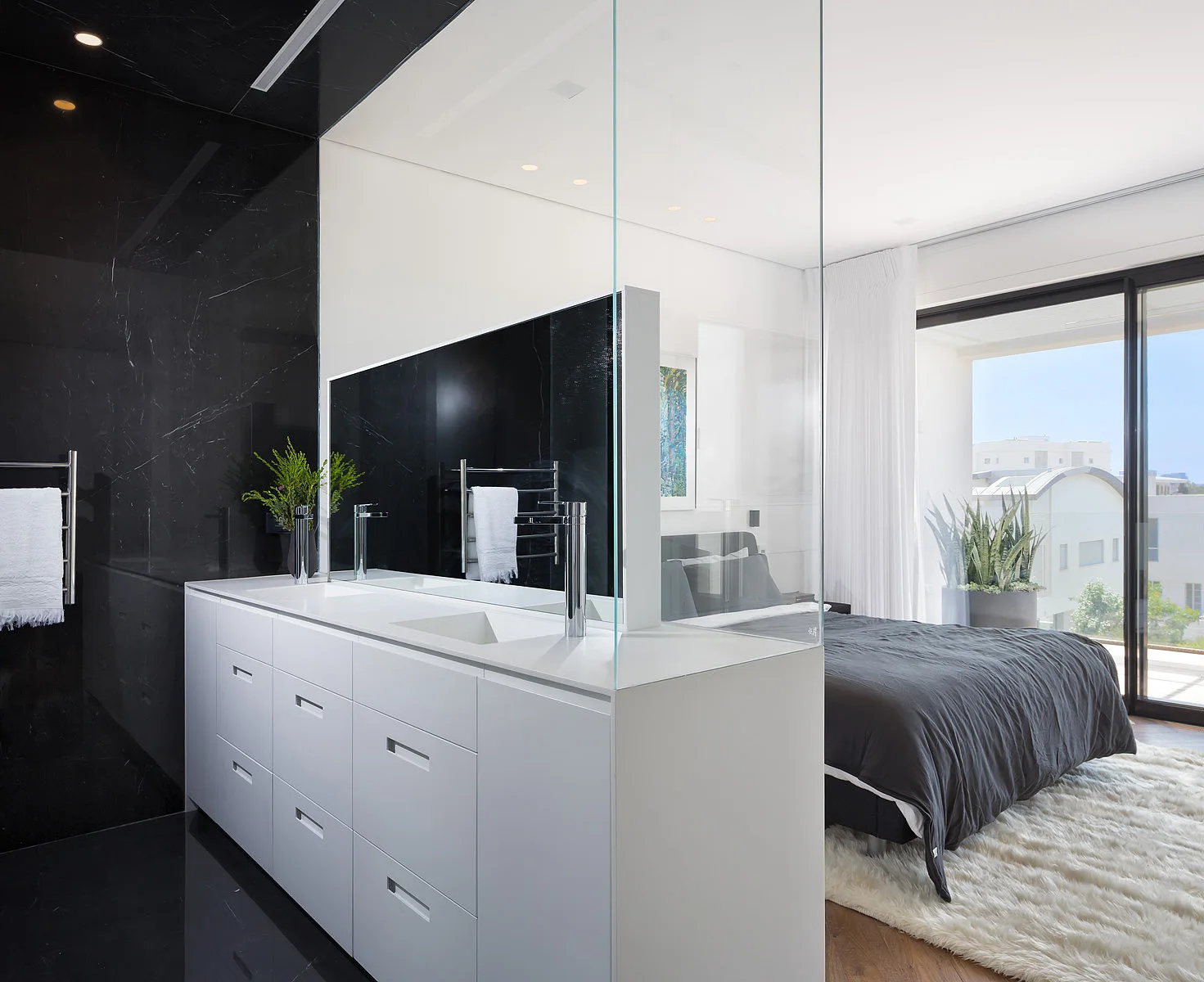 A white bathroom vanity and a tall glass partition divide a black-walled bathroom from a bedroom with a dark bed.