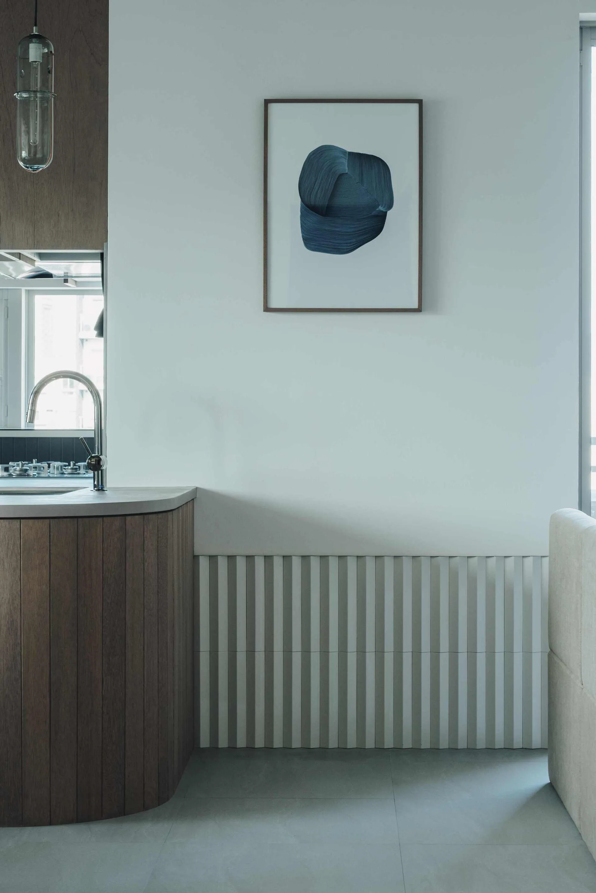 A modern kitchen space featuring a curved wood cabinetry island with a grey countertop, white fluted wall paneling, and a framed blue abstract art print.