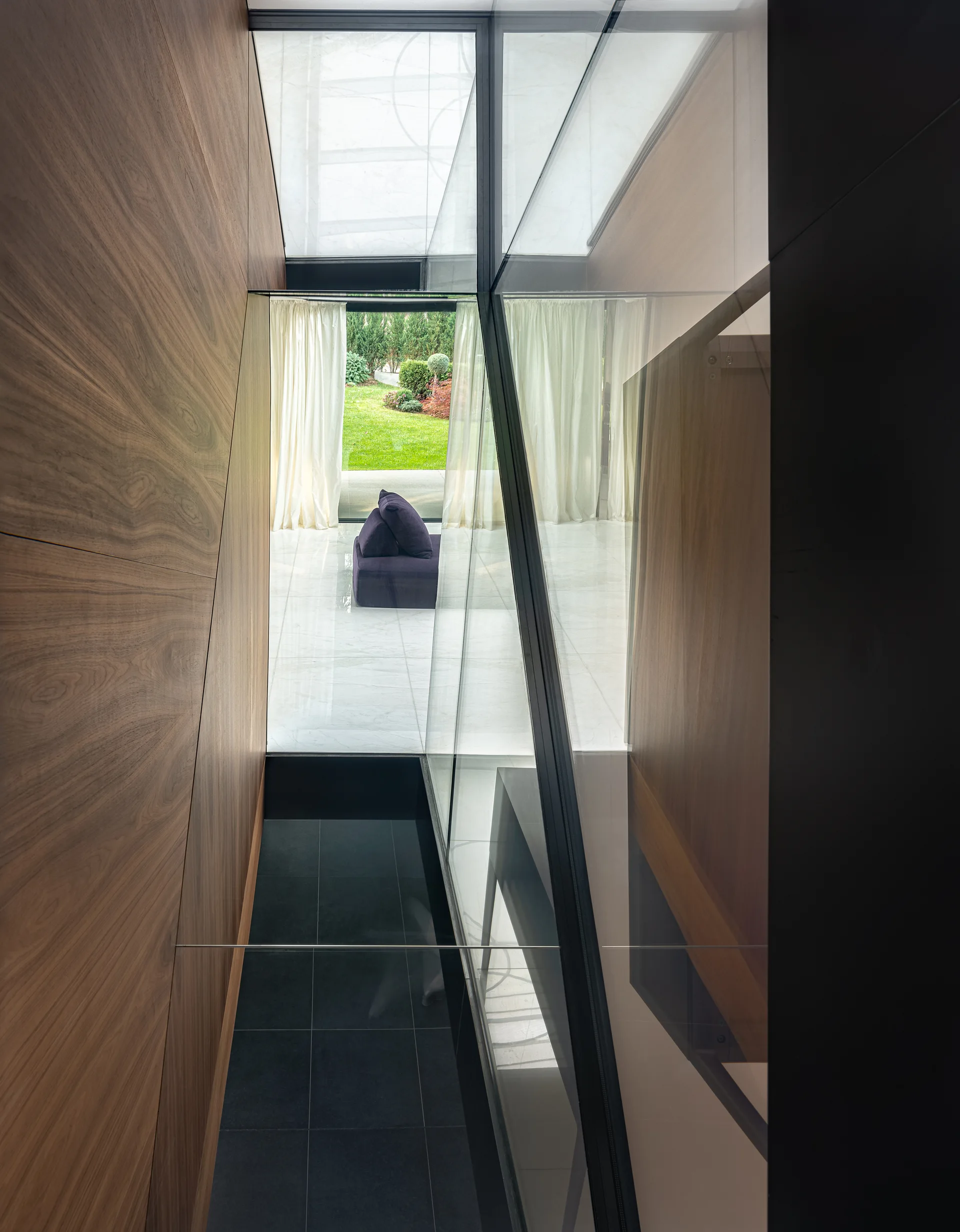 A hallway with warm wood-veneered walls and a glass partition looks into a bright white living room featuring a single purple sofa and a garden view.