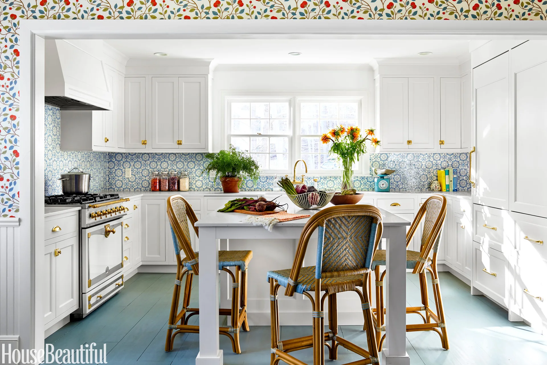 A white kitchen features a central island with woven bistro stools, painted blue wood floors, and a blue patterned tile backsplash.