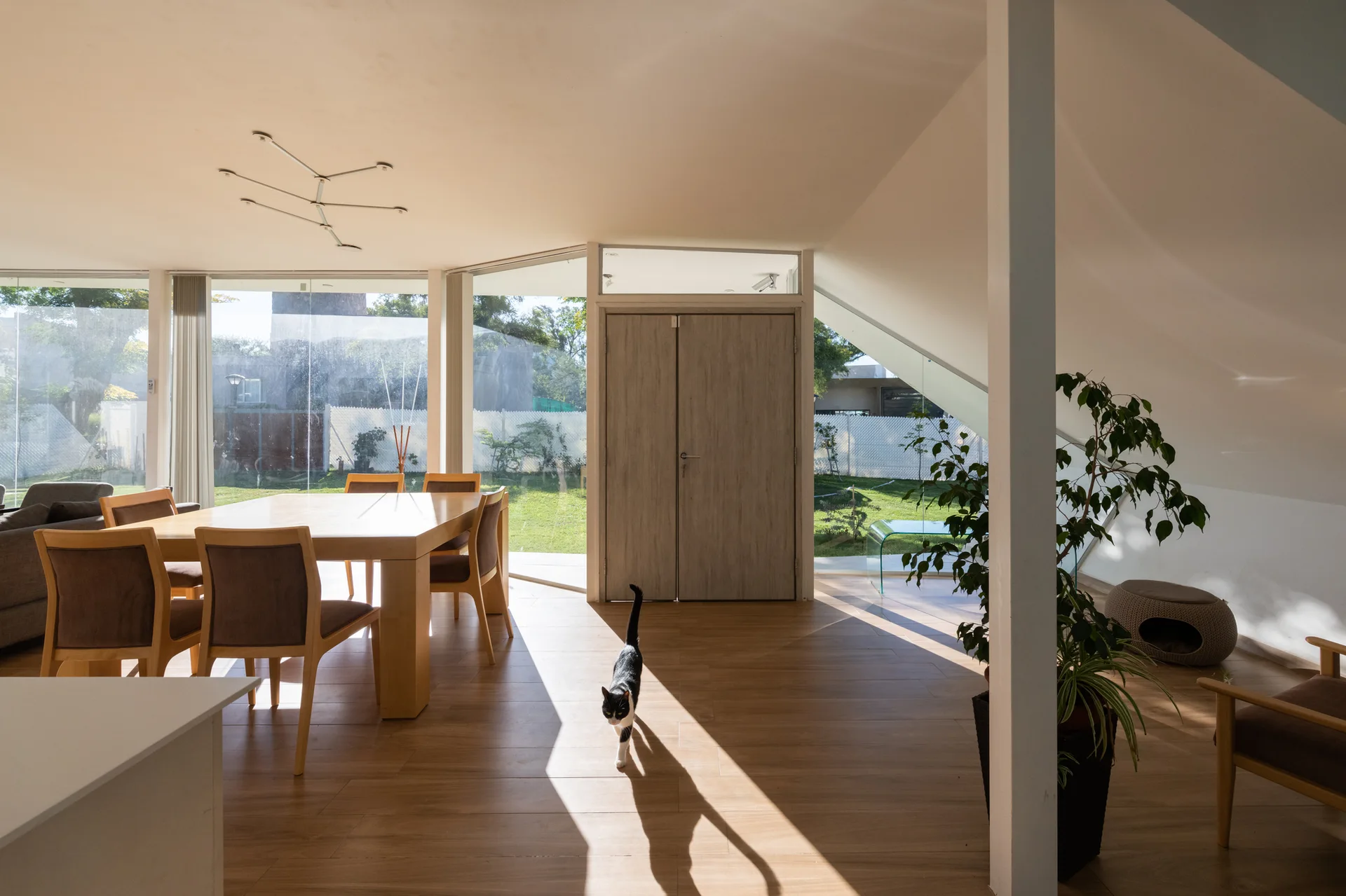 A modern dining room features a light wood table, chairs, a sloped white ceiling, and expansive floor-to-ceiling windows looking out onto a green yard.