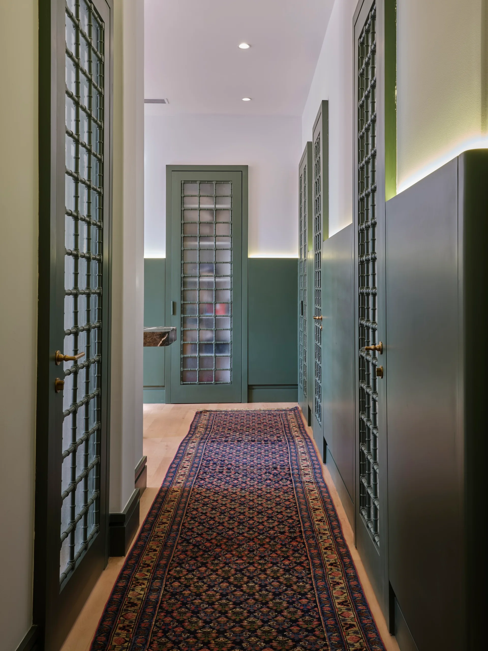 A narrow hallway featuring sage green paneled wainscoting, doors with decorative metal grilles, a patterned runner rug, and glowing cove lighting.
