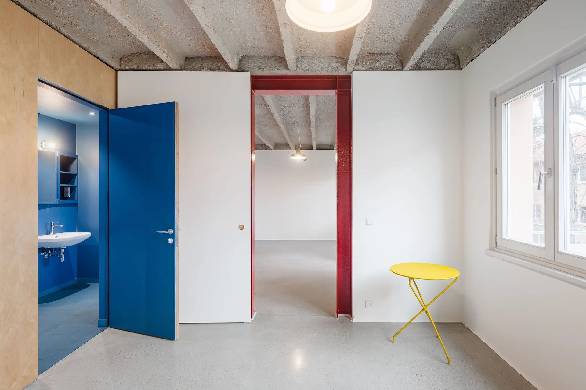 A minimalist white room features an exposed concrete ceiling, a bright blue bathroom door, a red steel entryway frame, and a yellow side table.