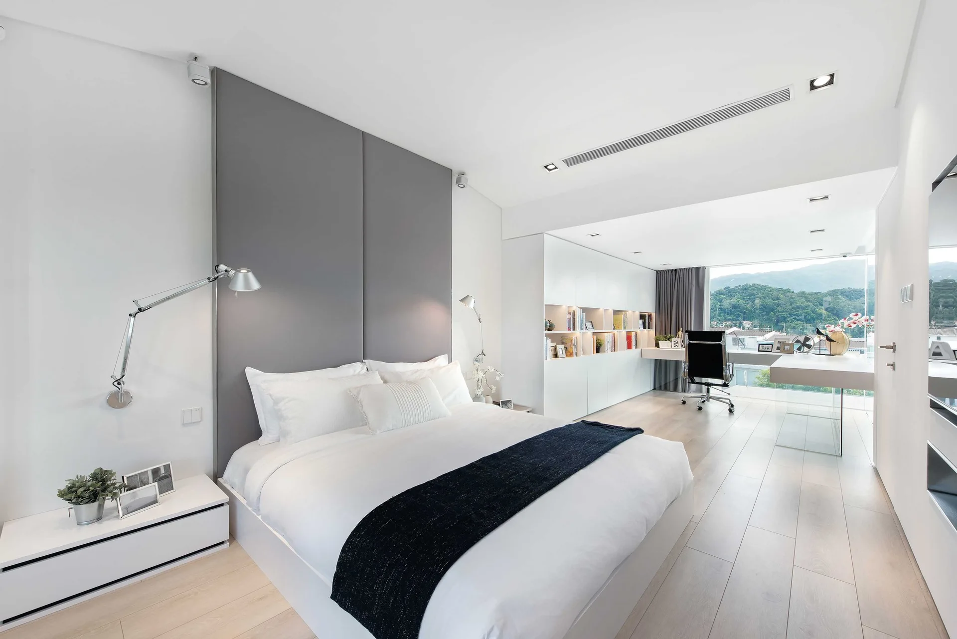 A modern white bedroom features a bed with a tall grey headboard, a built-in white desk with an office chair, and a large window.